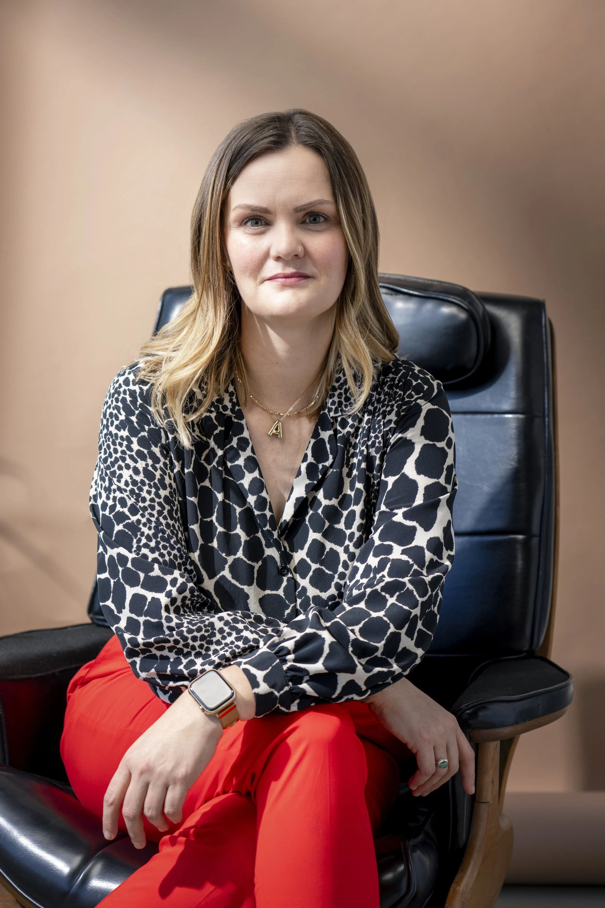 Amanda sits in a black leather chair with her legs crossed looking at the camera with a smile. She is wearing a black blouse and red pants.