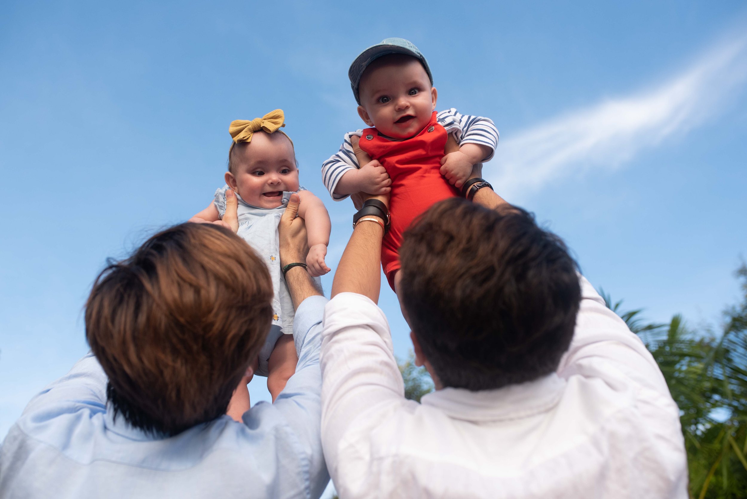 Deux hommes LGBTQ+, Gustavo et Romain, soulevant leurs deux bébés nés par GPA aux États-Unis.