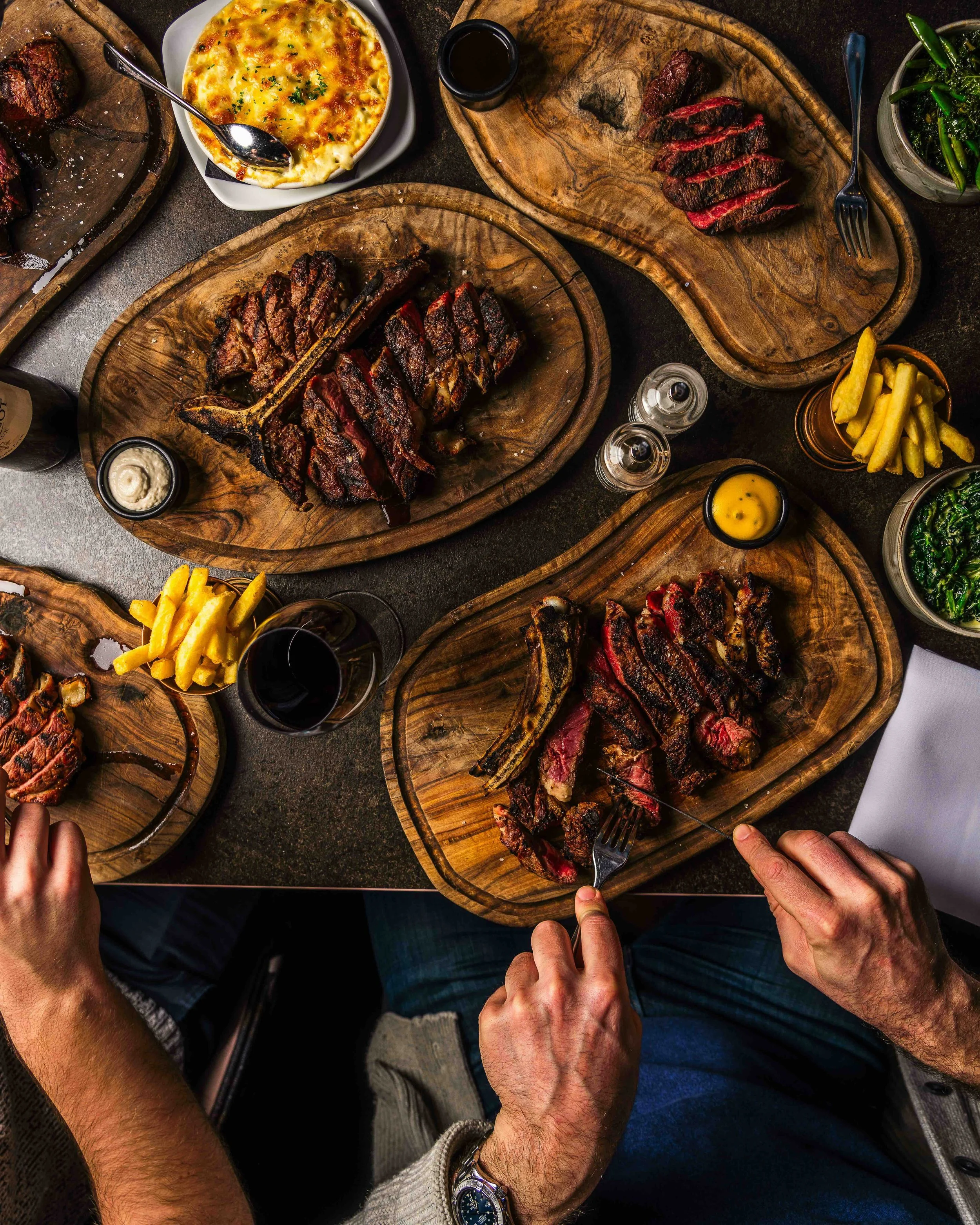 Sliced steaks on wooden boards, accompanied by fries, wine, gratin dish, and sautéed greens on a table.