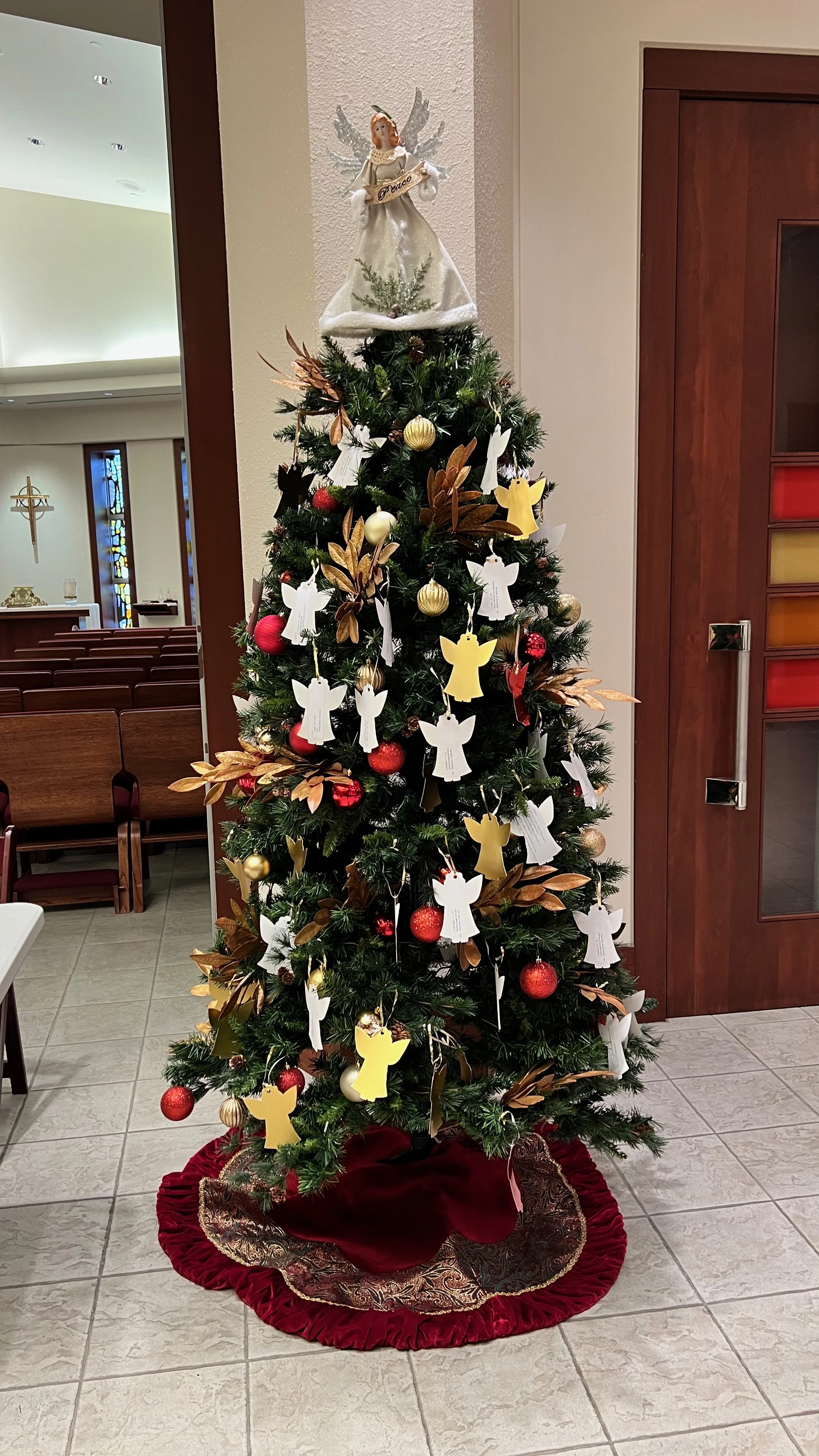 Decorated Christmas tree with gold and red ornaments, paper angels, and an angel topper holding a 'Peace' sign, placed on a red and gold tree skirt inside a church.