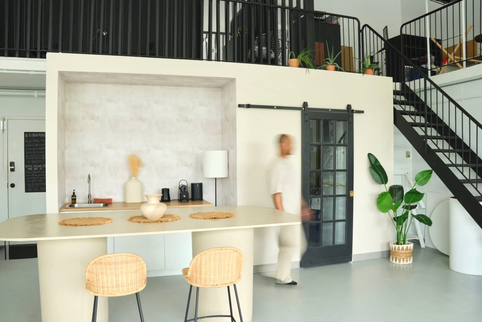 Intérieur d'une cuisine moderne avec un comptoir beige, deux chaises en bois, et des décorations en céramique. Un homme marche près d'une porte vitrée noire, avec une plante verte à côté.
