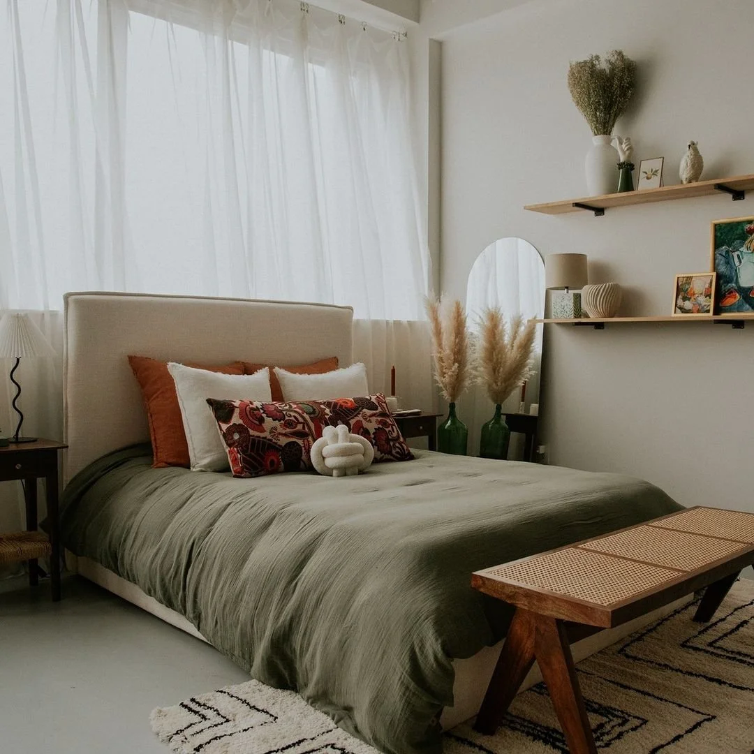 Chambre à coucher avec lit tapissé de draps verts, plusieurs coussins décoratifs, étagères avec vases et décorations, plantes pampas en verre, miroir rond, et une table en bois.