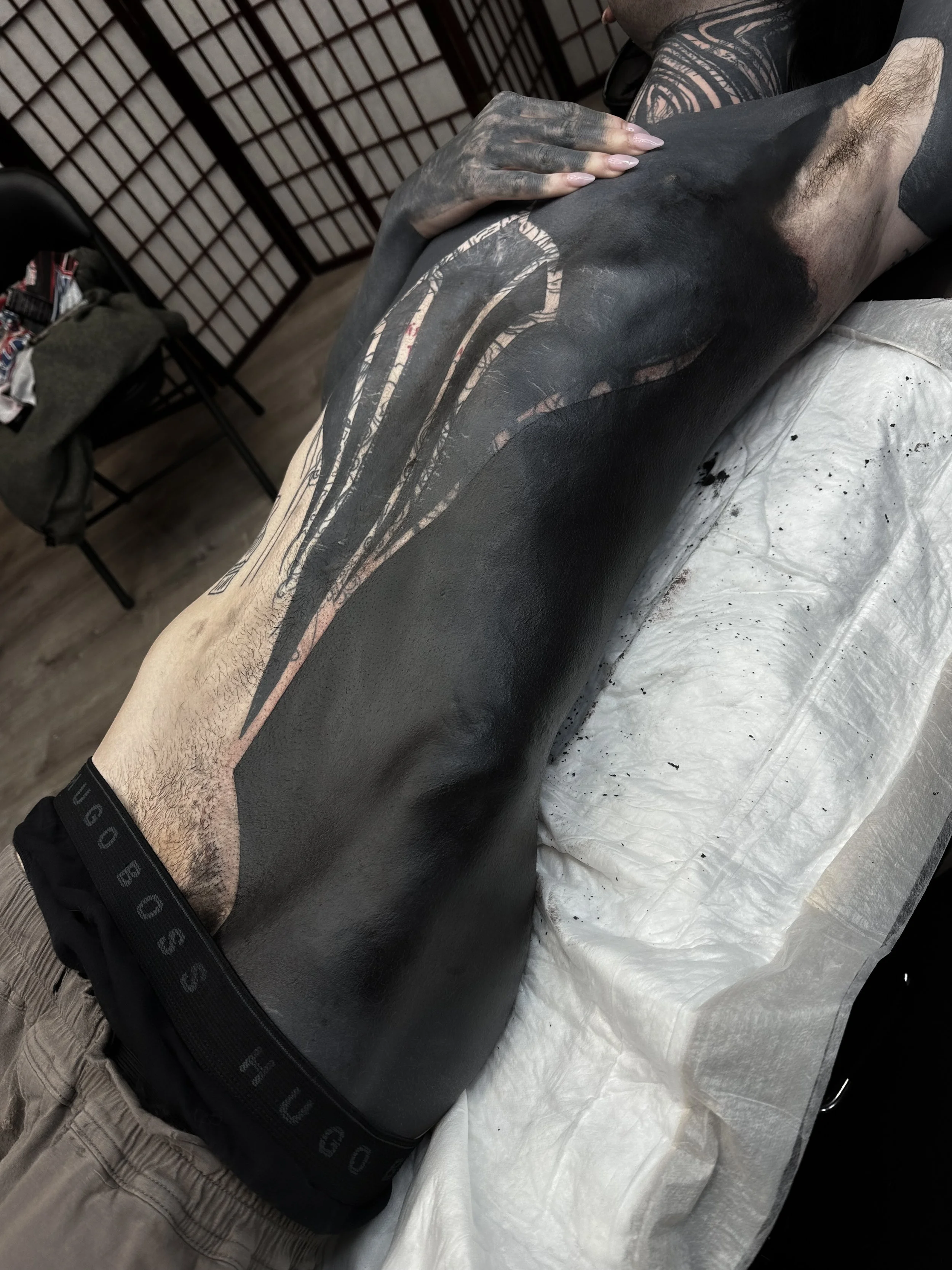 A person receiving a tattoo on their thigh in a tattoo studio. The tattoo artist's hand is resting on the person's thigh.
