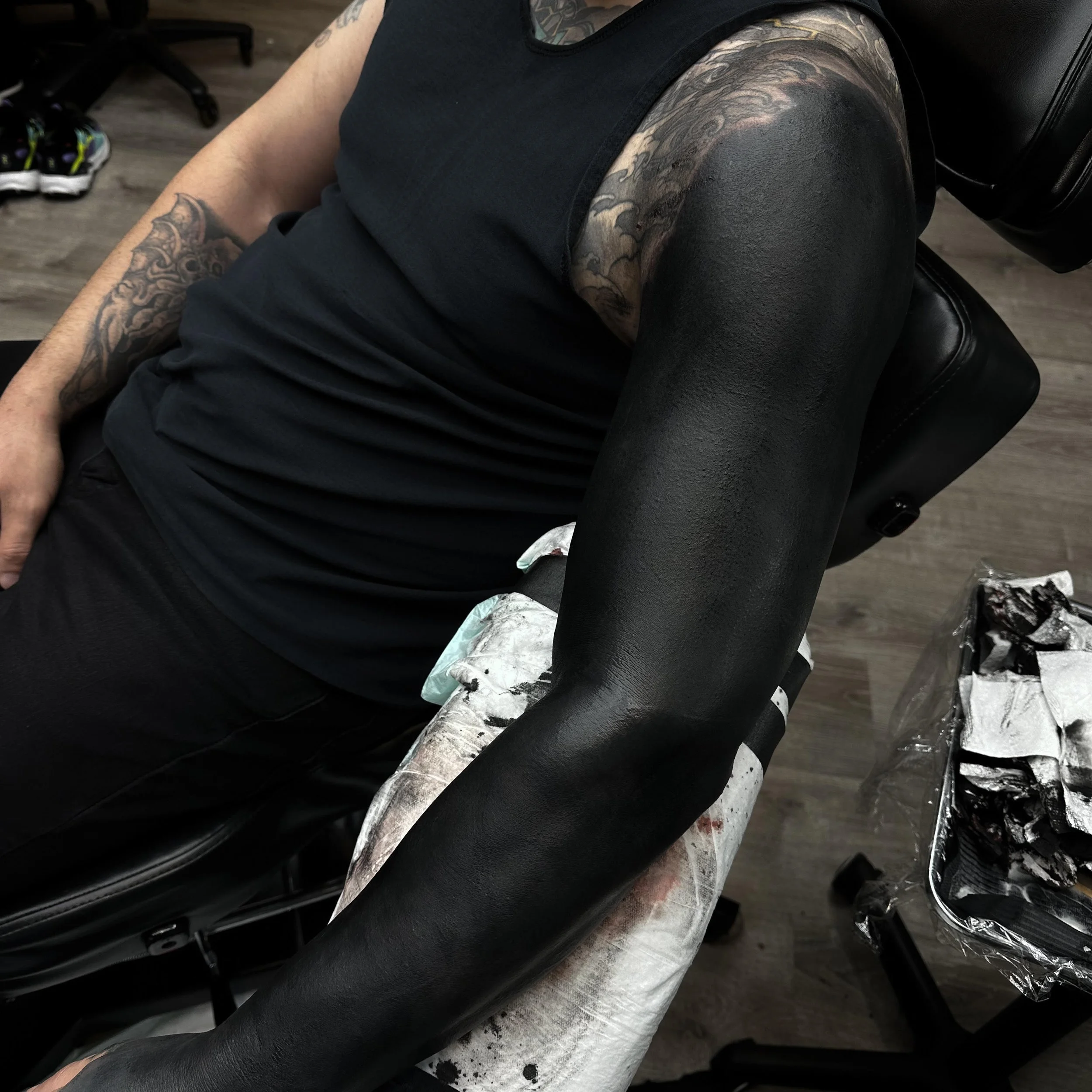 Man lying on a tattoo chair with freshly inked black sleeve tattoo on his right arm and colorful tattoo on his shoulder.
