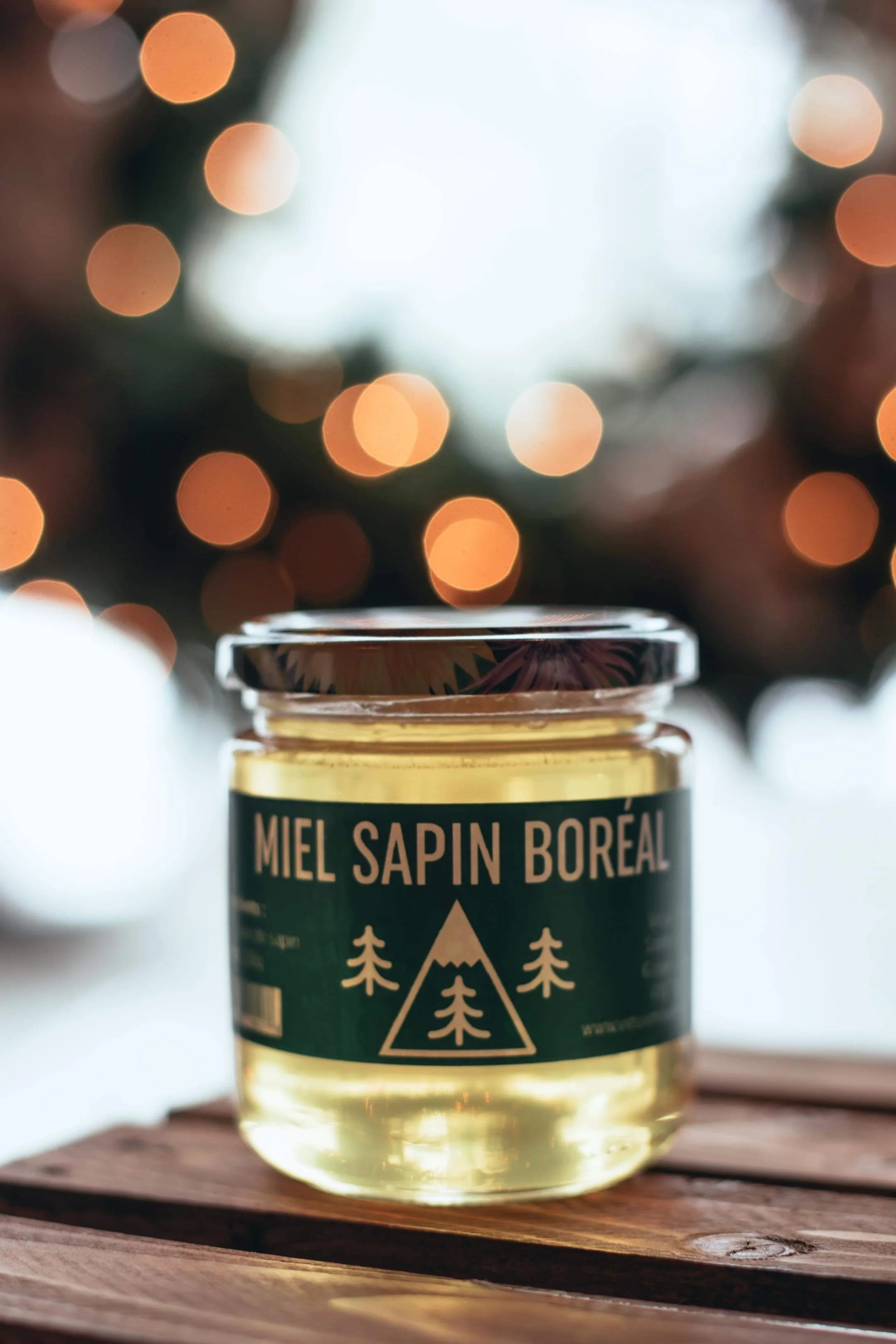 Pot de miel nommé "Miel Sapin Boréal" sur une table en bois, fond flou de lumières lumineuses ambiance chaleureuse et festive.