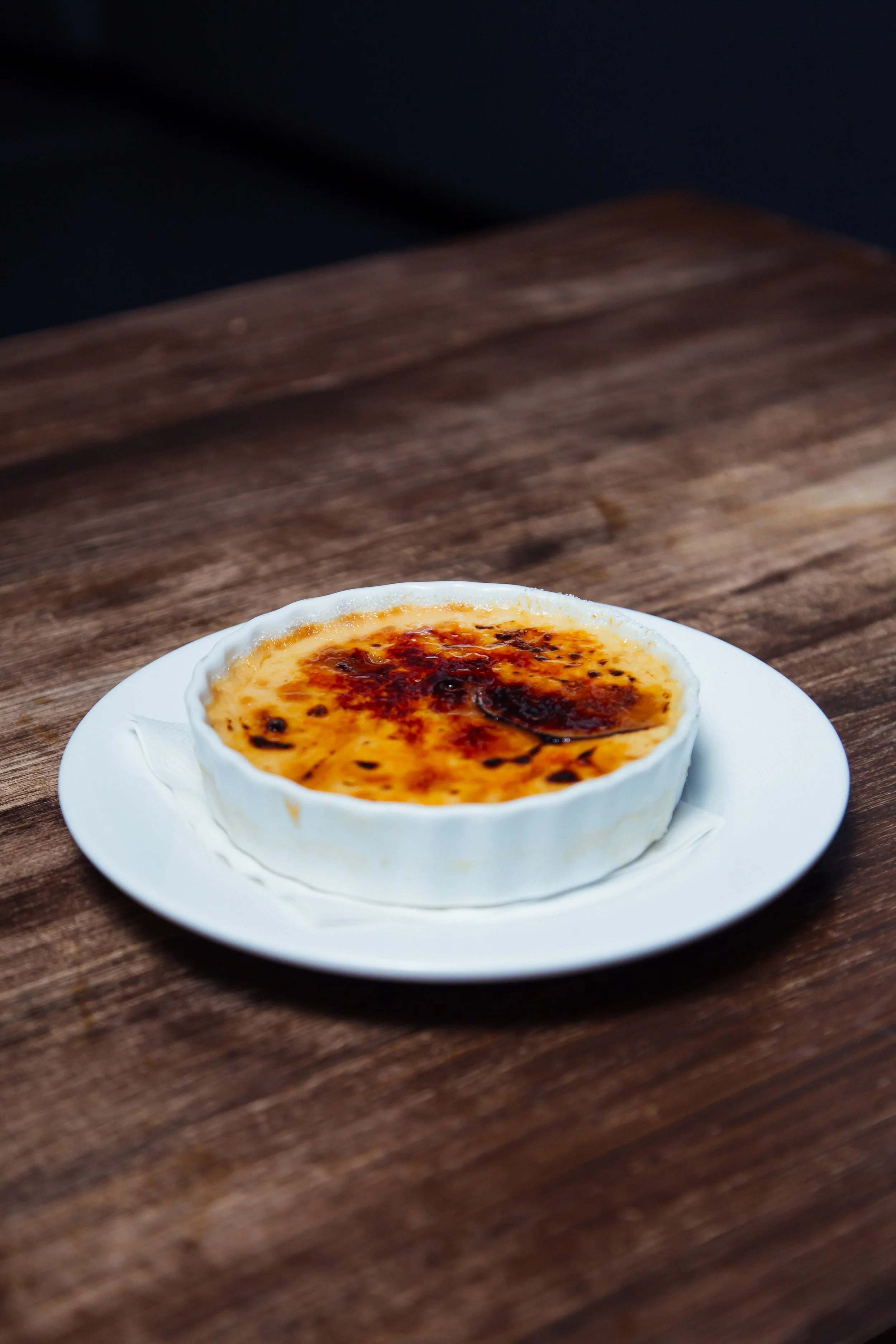 Crème brûlée dans un ramequin blanc sur une assiette blanche, posée sur une table en bois.