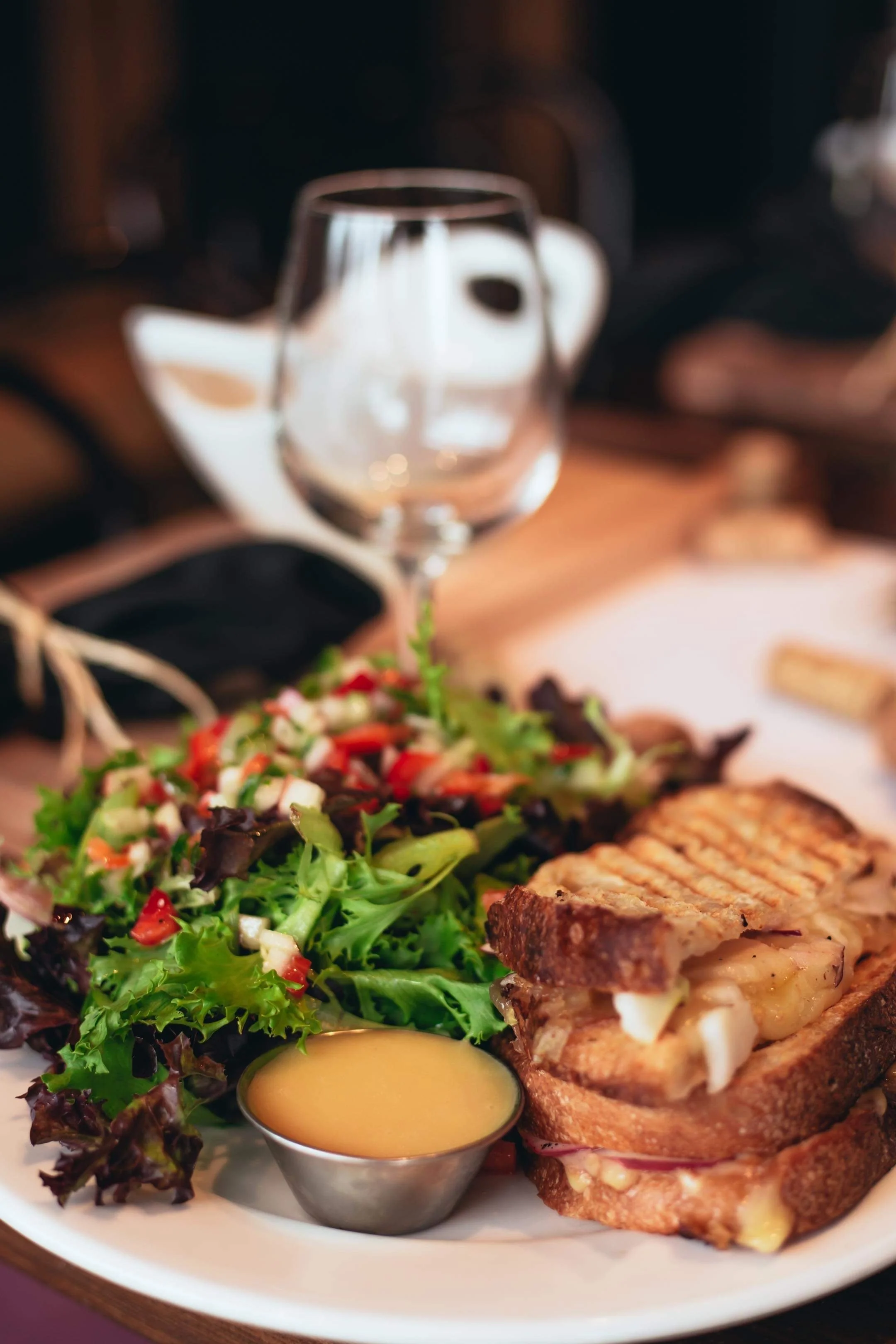 Assiette avec salade verte, sandwich au pain grillé avec fromage, accompagnée d'une petite coupe de sauce et d'un verre de vin vide.