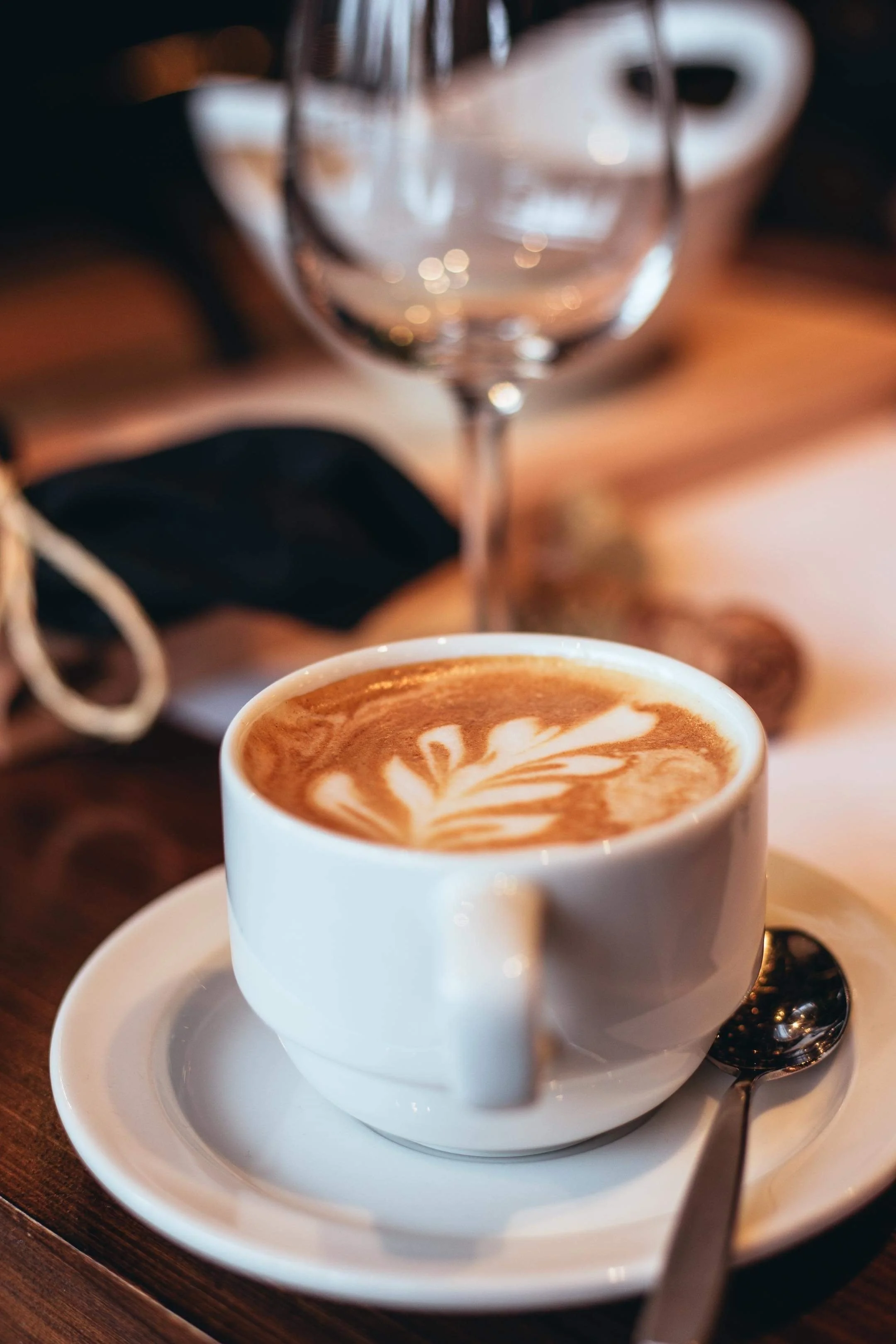 Tasse de café latte avec dessin en forme de feuille, accompagnée d'une cuillère en métal, avec un verre d'eau en arrière-plan, sur une table en bois.