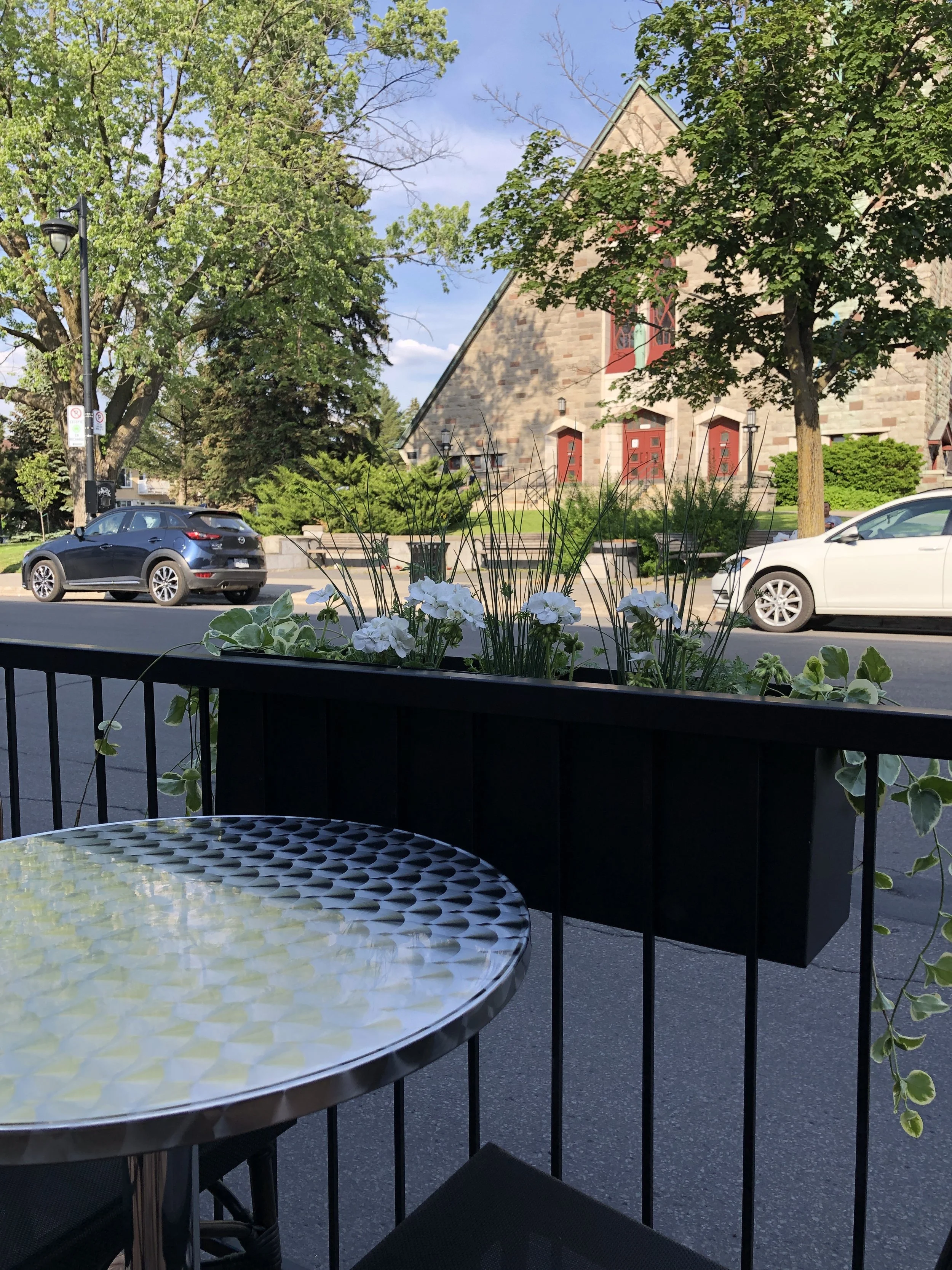 Une terrasse de café avec une table en métal, une barrière en métal avec une jardinière de fleurs blanches et vertes. En arrière-plan, une rue avec des voitures et un bâtiment en pierre avec des fenêtres rouges, entouré d'arbres verts.