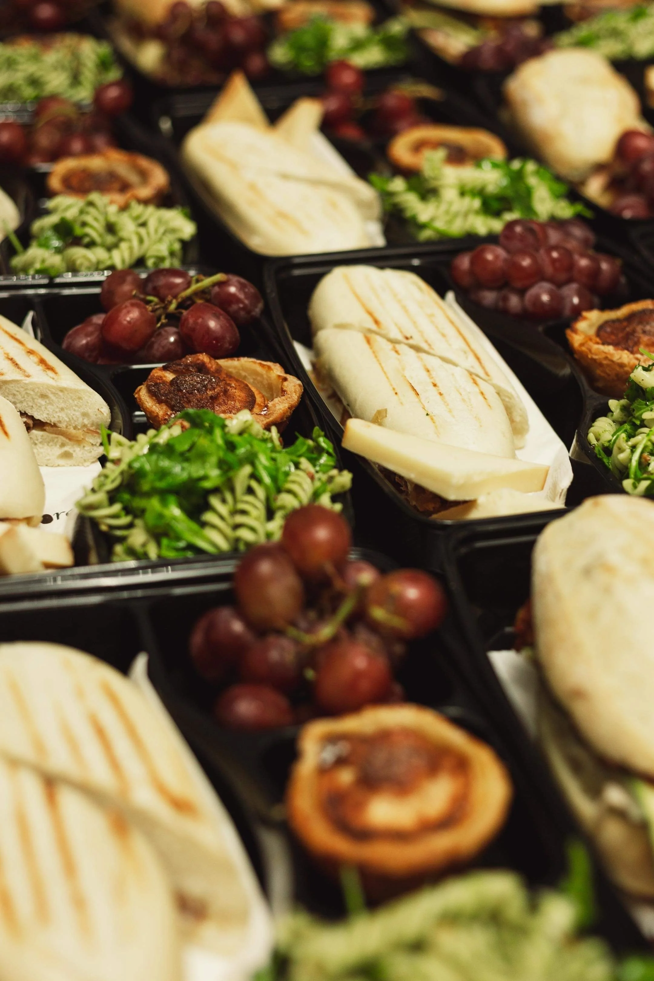 Assortiment de sandwiches, raisins rouges, pâtes fusilli avec des légumes verts, et petits quiches dans des plateaux de repas.