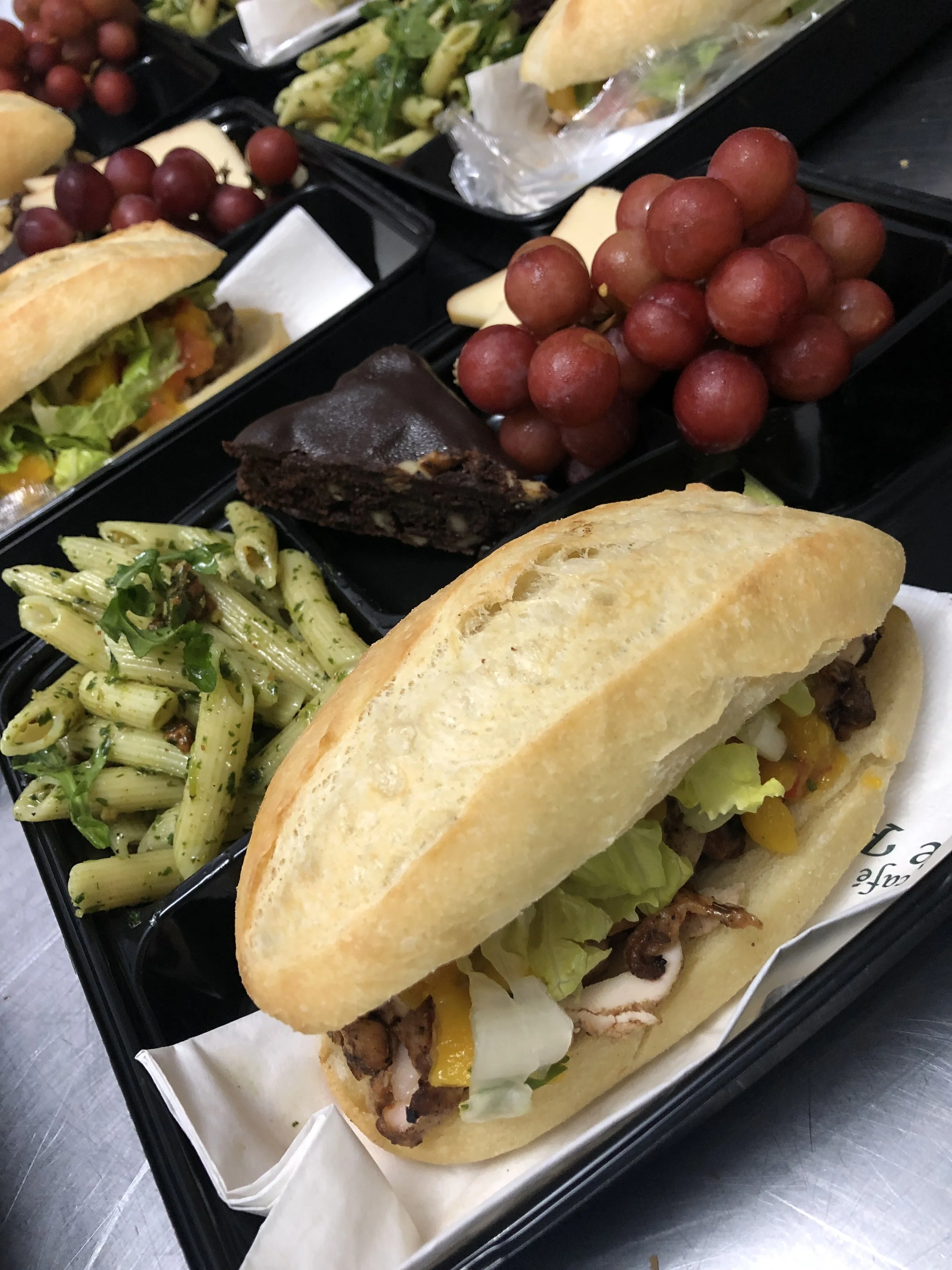 Un plateau avec un sandwich au pain baguette, de la salade, du poulet, et des légumes, accompagné de pâtes, de raisins rouges, un morceau de gâteau au chocolat, et des légumes verts.