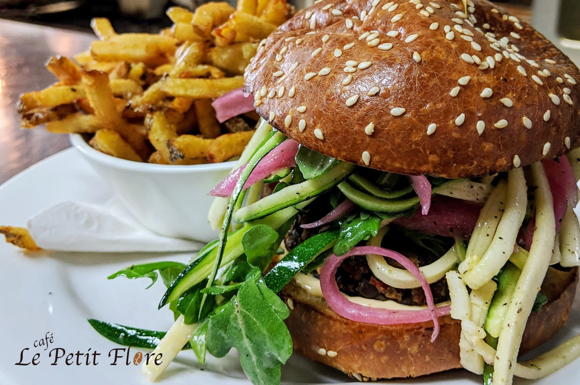 Sandwich burger avec steak haché, légumes frais, oignons rouges, concombre, et pain brioché avec des graines de sésame, accompagné de frites maison, servi sur une assiette blanche