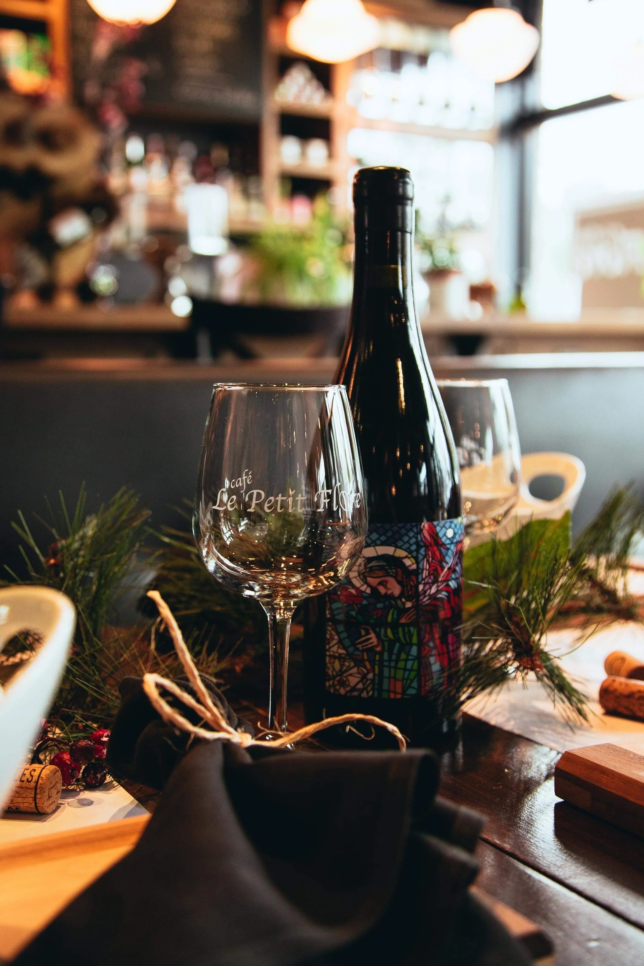 Une bouteille de vin avec une étiquette colorée, un verre à vin vide, décorations de sapin, et éléments de cuisine dans un restaurant, ambiance chaleureuse.