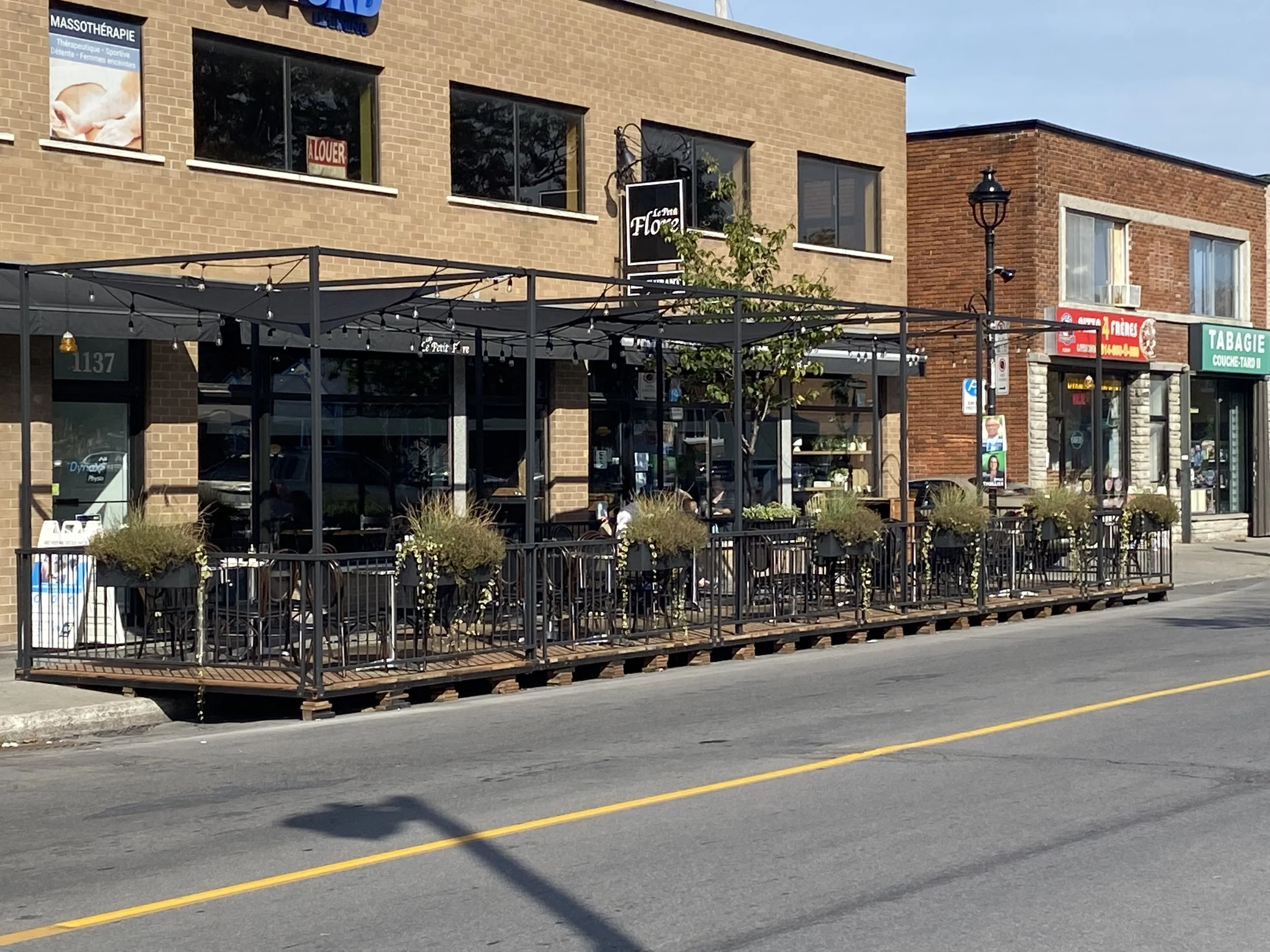 Terrasse extérieure d'un café ou restaurant avec des plantes en pot et des tables, située sur un trottoir devant des bâtiments commerciaux. Le ciel est clair et il fait jour.