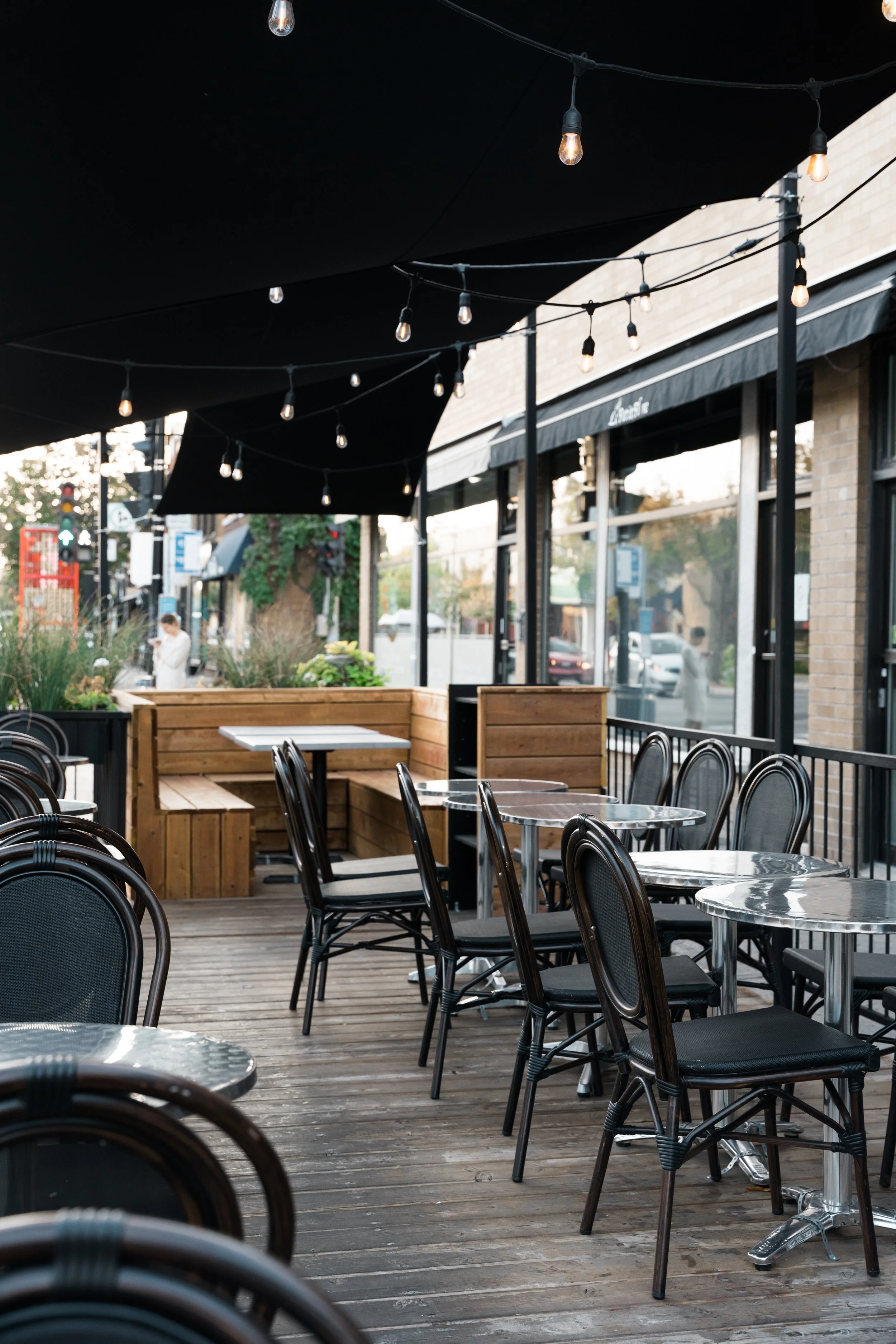 Terrasse du café Le Petit Flore avec tables en métal, chaises en bois, toit noir avec guirlandes lumineuses, vitres avec cadre noir, ambiance extérieure