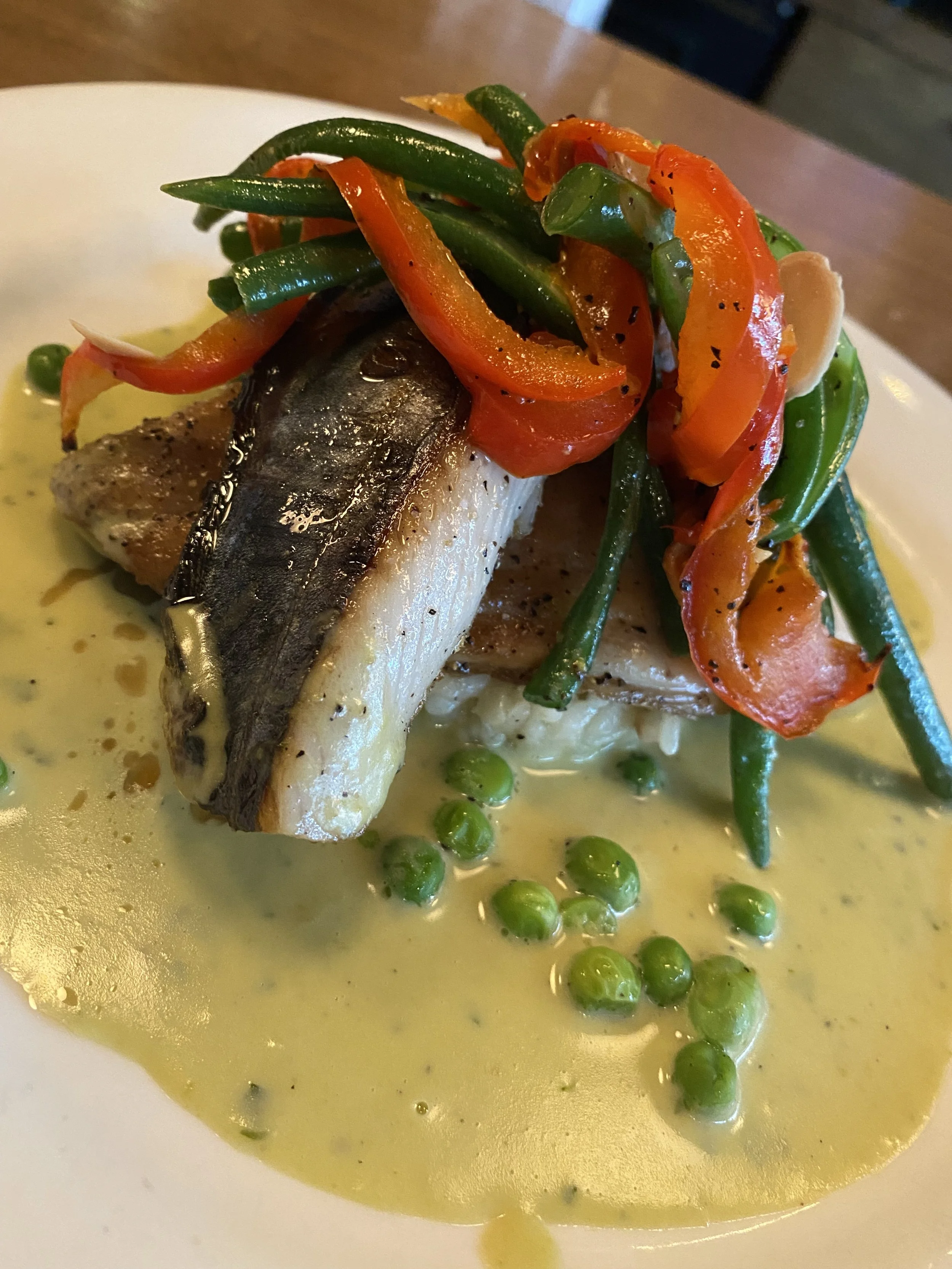 Poisson grillé avec légumes sautés, pois verts, et sauce crémeuse sur une assiette blanche.