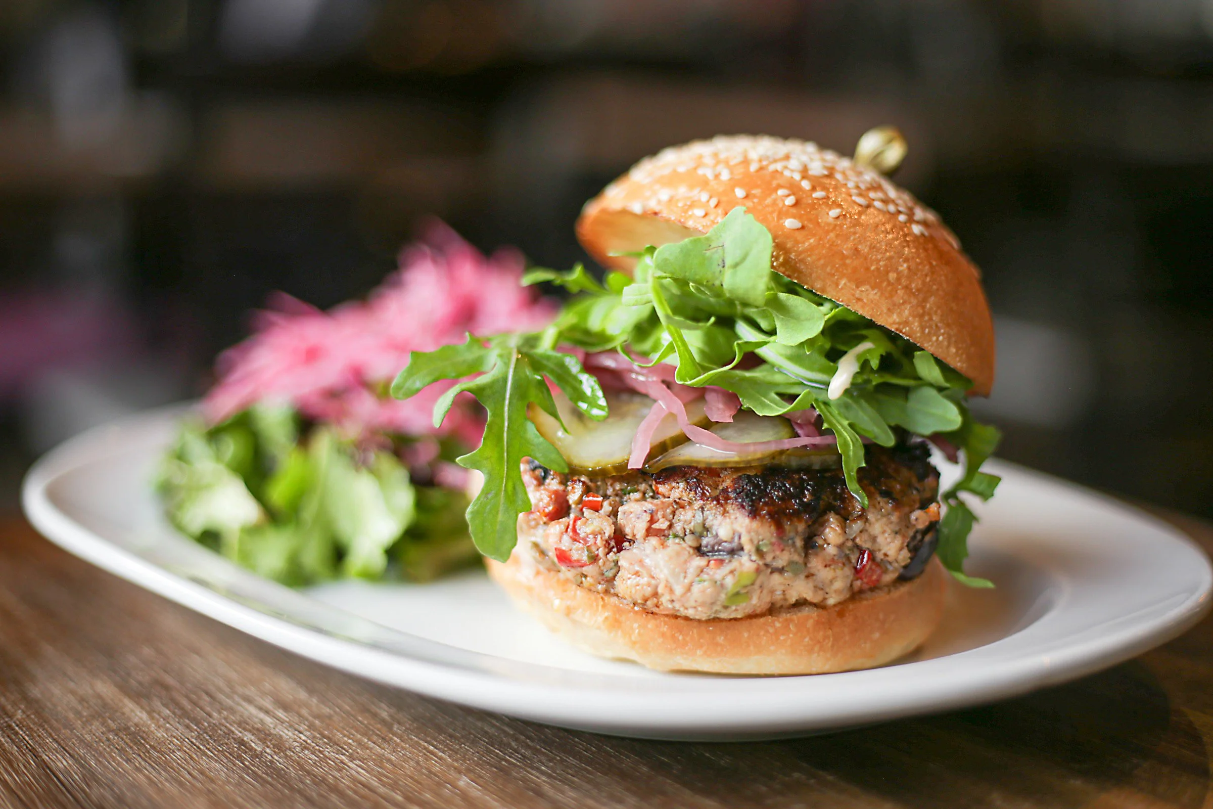 Burger avec galette de viande, salade, cornichon, oignons rouges dans un pain au sésame, accompagnée de salade verte et fleurs roses.
