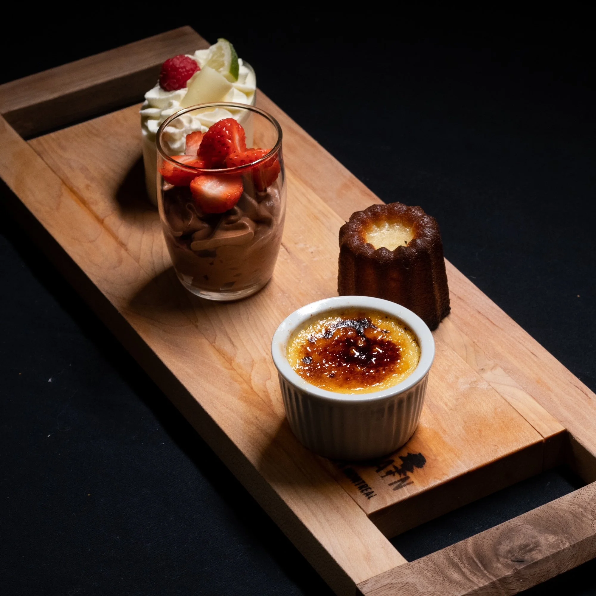 Assortiment de desserts sur une planche en bois, comprenant une crème brûlée, un gâteau coulant, un verre de mousse au chocolat aux fraises, et une tartelette
