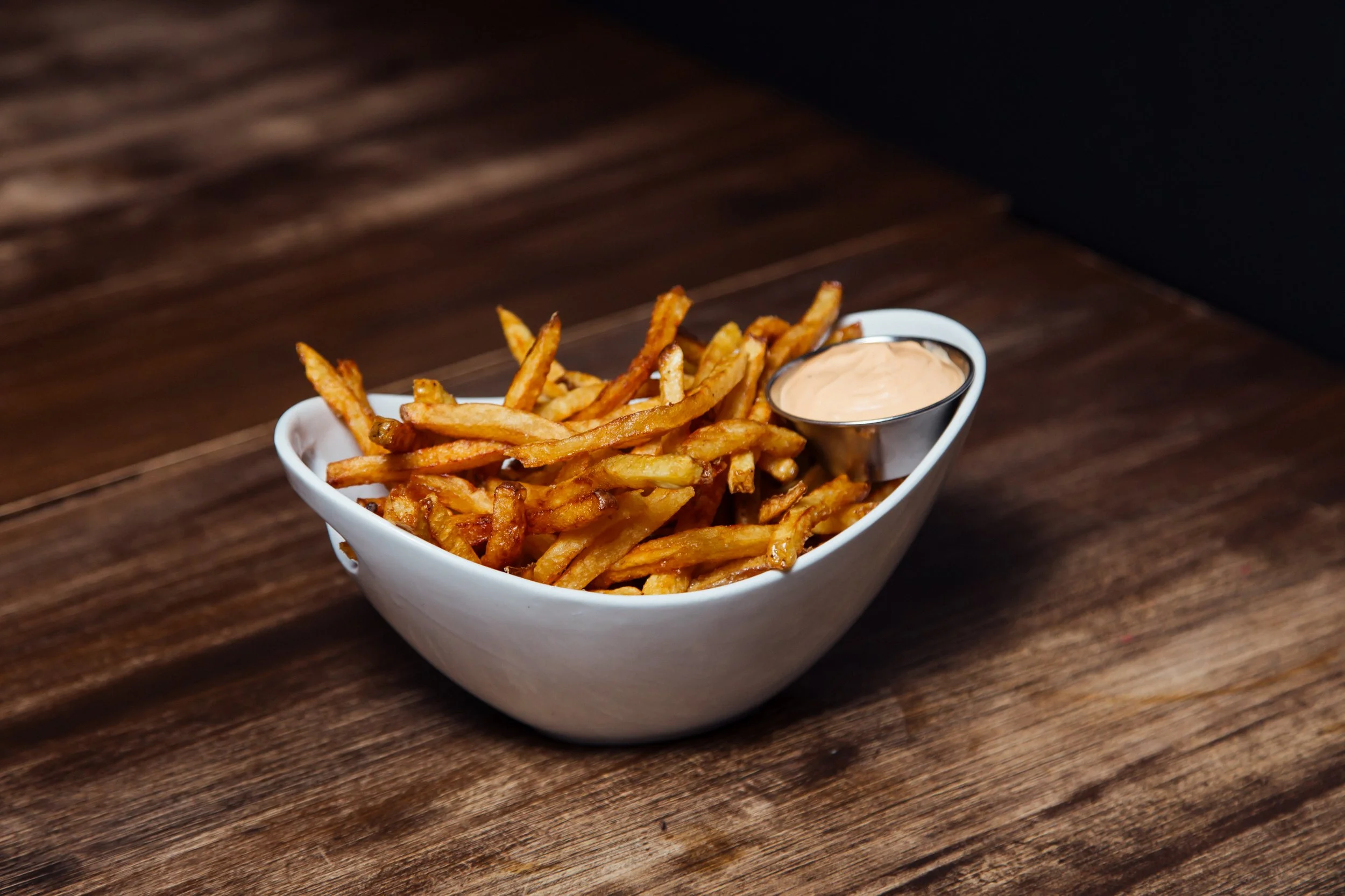 Frites dorées dans un bol blanc avec une petite coupe de sauce rose, posés sur une table en bois.