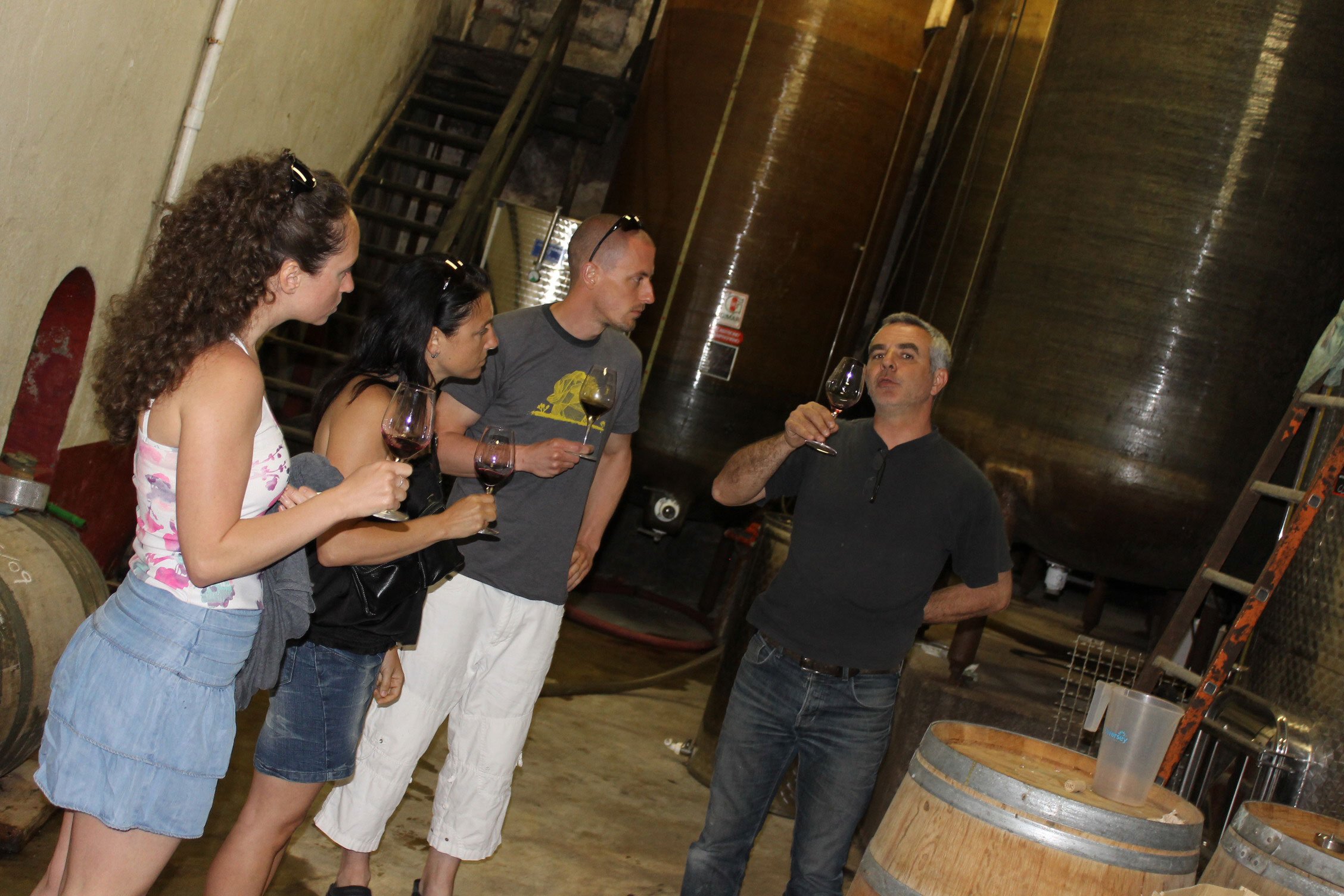 Groupe de quatre personnes dans une cave à vin, tenant des verres de vin rouge, écoutant un homme qui explique, avec de grandes cuves en arrière-plan.