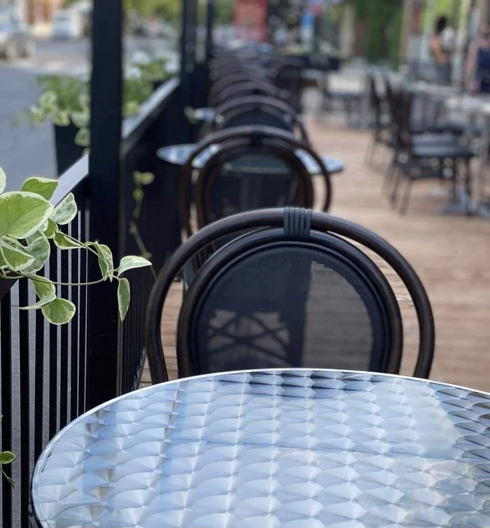 Une terrasse de café vide avec une table en verre et des chaises en rotin en plein air, avec un peu de végétation à gauche.