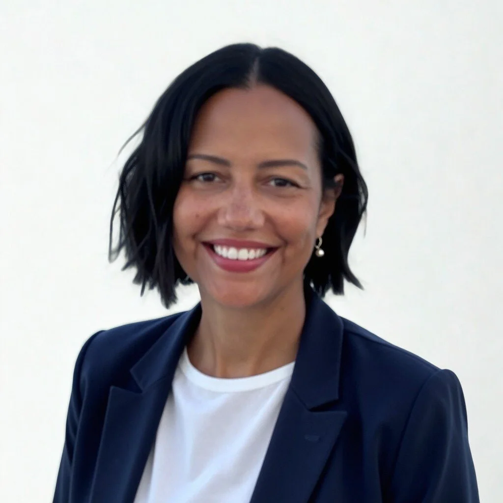 Portrait of a woman with long dark hair, wearing a blue blazer and gold buttons, smiling outdoors with a cloudy sky and water in the background.