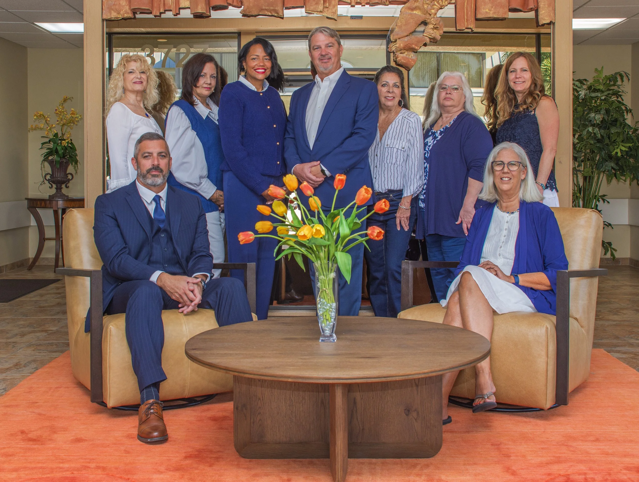 A group of ten diverse professionals, dressed in business casual attire, pose together in an office lobby with a large glass window, potted plants, and a wooden decor element in the background. They are gathered around a coffee table with a vase of orange and yellow tulips, seated on beige leather chairs and standing behind.