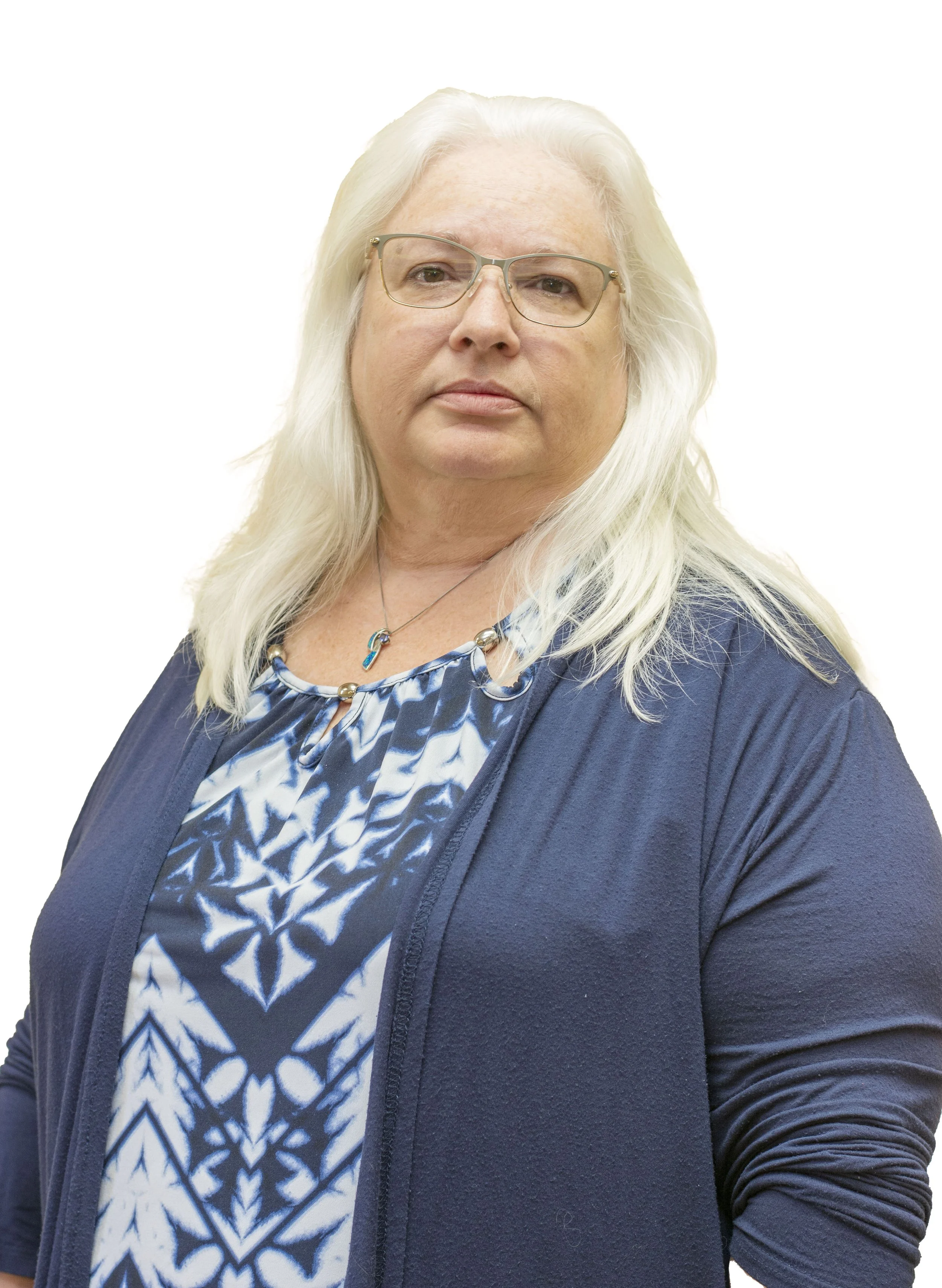 A smiling woman standing outdoors near a body of water, with palm trees, a cloudy sky, and a railing in the background. She is wearing a blue checked top with a white undershirt and a silver chain necklace with a heart-shaped pendant.