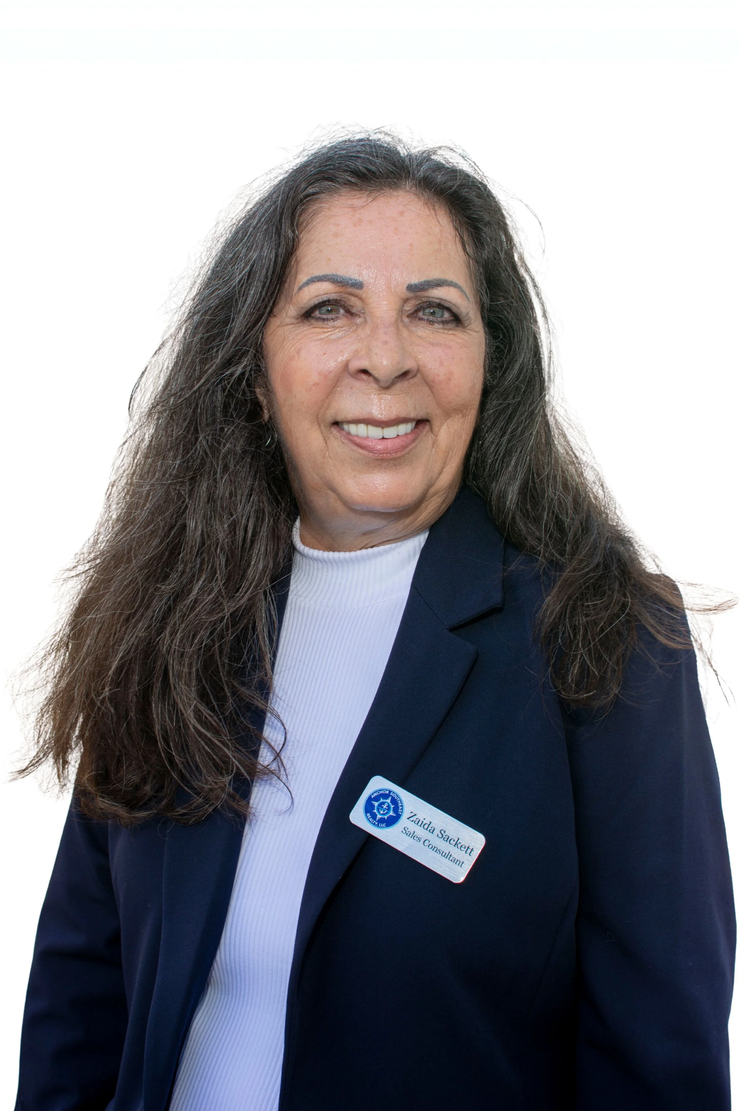 A woman outdoors near water, smiling, wearing a navy blazer with a name tag that reads Zaida Sackett, Sales Consultant.