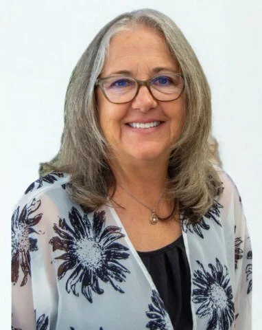 A smiling middle-aged woman with shoulder-length gray hair and glasses, wearing a white blazer with blue floral patterns over a black top, and a small necklace.