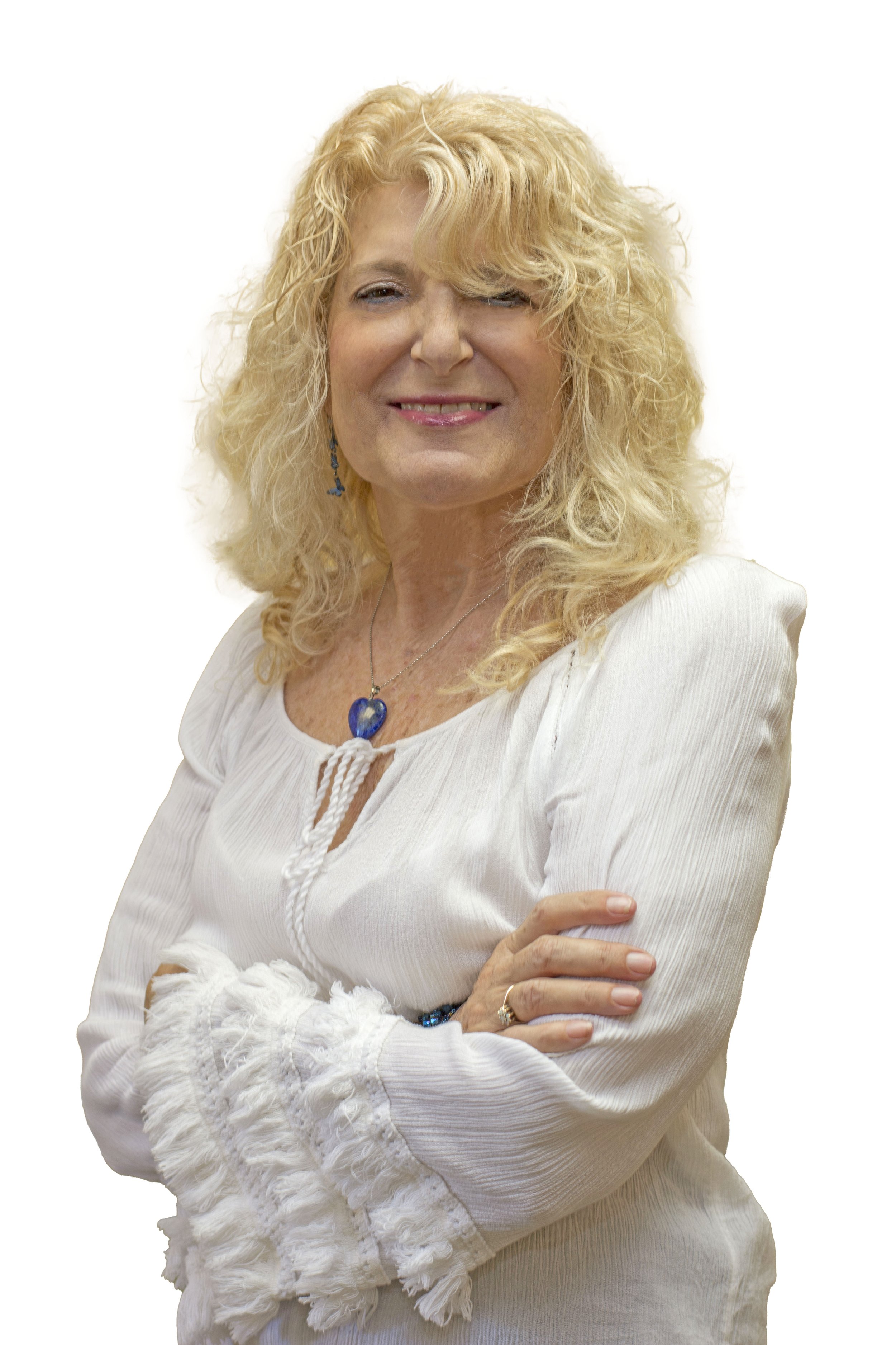 A smiling woman with blonde curly hair standing outdoors near a body of water, wearing a white sleeveless top, large blue earrings, and a blue beaded necklace.
