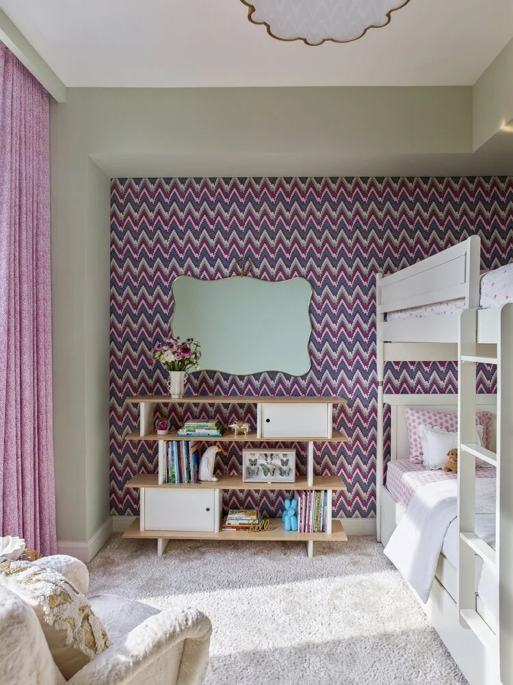 Children's bedroom with patterned wallpaper, white bunk bed, and bookshelf with books and decor accents. Features pink curtains, a beige armchair, and decorative elements on a plush carpet.