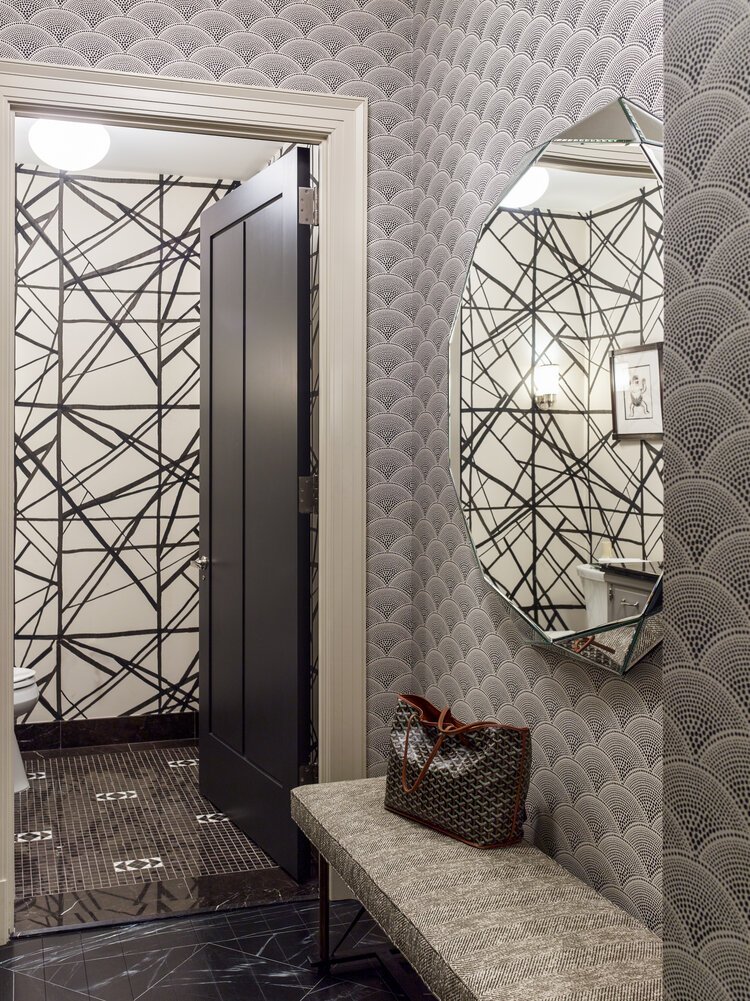 Modern bathroom with geometric wallpaper, a large round mirror, black door, and a bench with a tote bag.