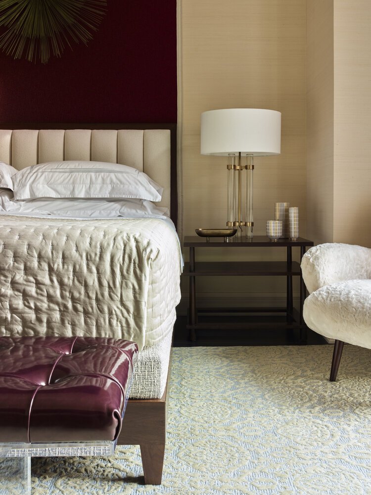 Modern bedroom interior with a beige upholstered bed, white linens, and a nightstand with a lamp. A woolly armchair and a tufted bench in maroon leather are visible, along with a patterned rug on the floor. A large accent piece is mounted on the wall