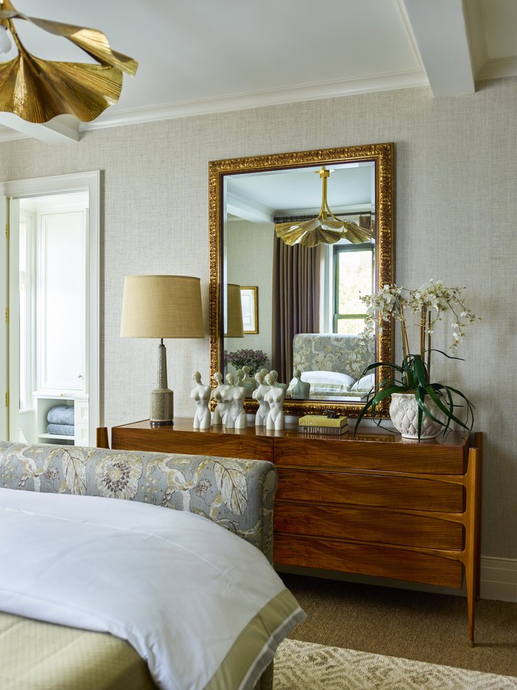 Bedroom interior with a large framed mirror, wooden dresser, decorative sculptures, table lamp, and a bed with floral-patterned headboard.