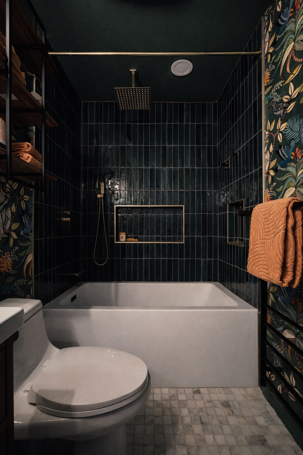 Modern bathroom with dark tiled shower area, white bathtub, and patterned wallpaper. Includes a toilet in the foreground, shelving with towels on the left, and an orange towel hanging on a rack on the right.