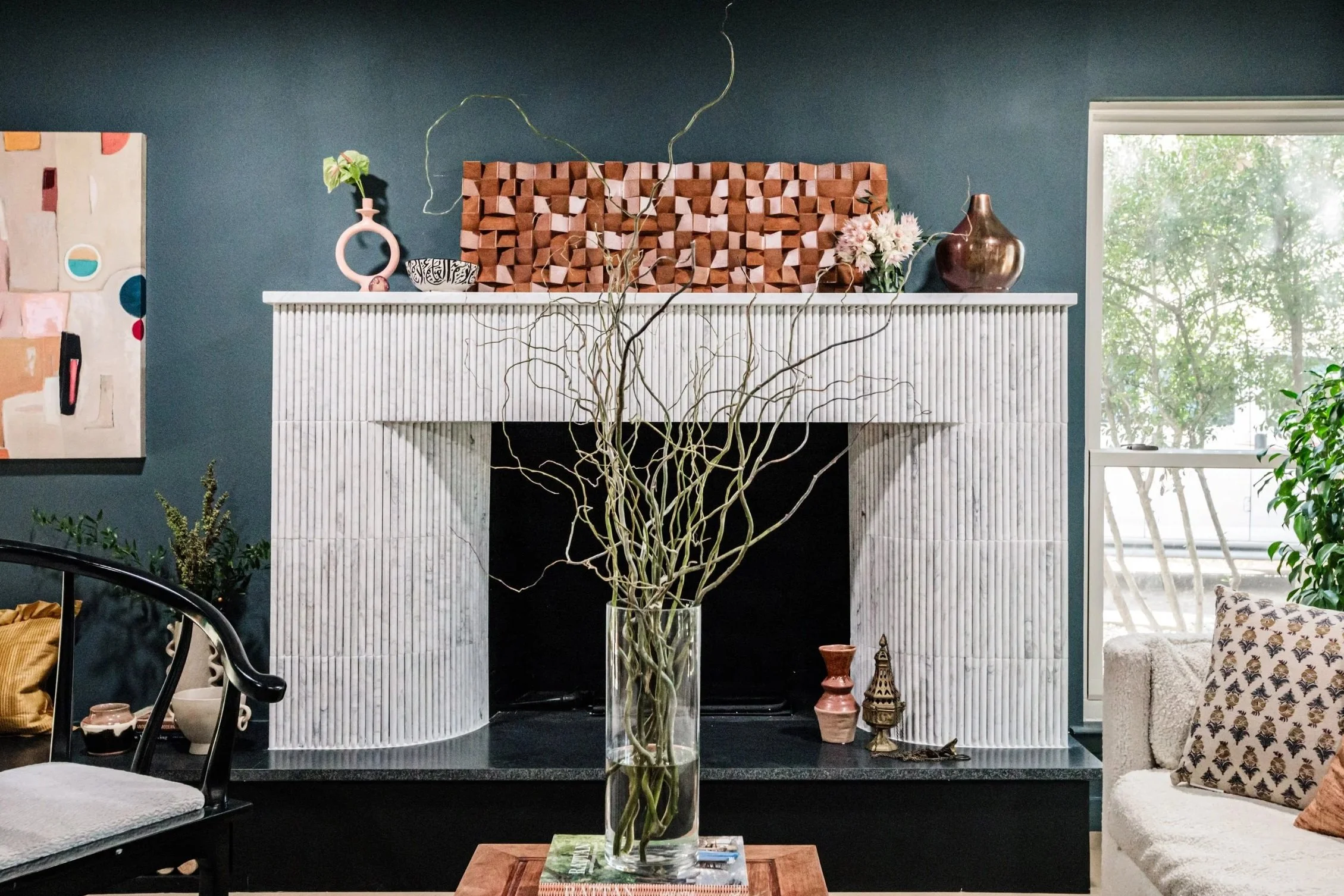 Living room with a white fireplace, a large glass vase with twisted branches on a wooden table in front, and decorative vases and flowers on the mantel. A window with greenery outside is on the right, and an abstract painting is on the left wall.
