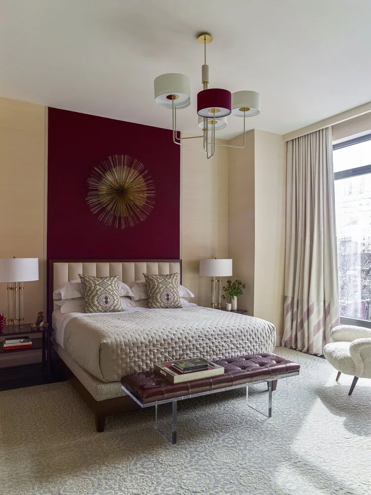 Elegant bedroom interior featuring a bed with a tufted headboard, patterned pillows, and a textured quilt. A burgundy accent wall with a decorative sunburst centerpiece is behind the bed. A modern chandelier hangs from the ceiling. The room includes 