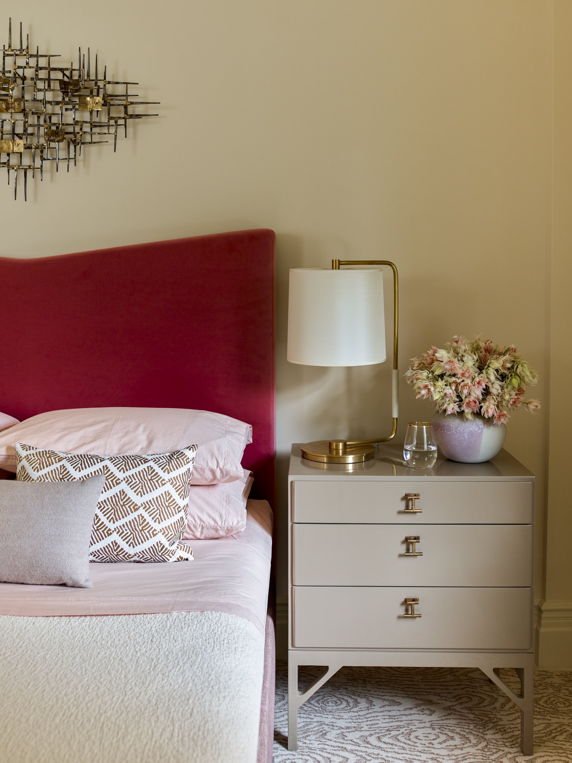Bedroom with a red velvet headboard, white and patterned pillows, beige nightstand, gold table lamp, and a vase of flowers.