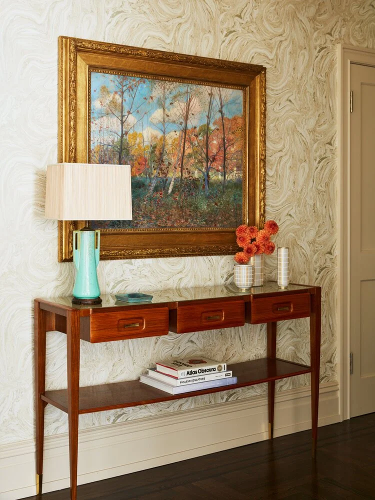 Elegant foyer with wooden console table, framed landscape painting, turquoise lamp, orange flowers in vase, and books underneath.