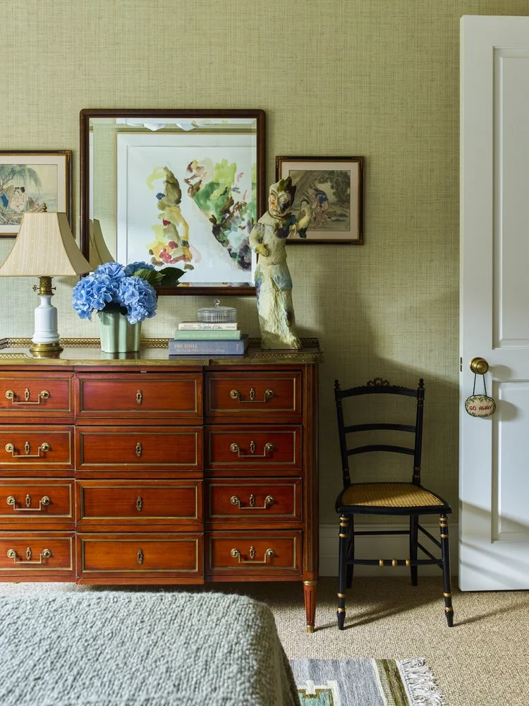 Bedroom interior with a wooden dresser, a table lamp, blue hydrangea bouquet, books, and a decorative statue. A mirror and wall art hang above the dresser. A black chair sits next to a door with a "do not disturb" sign.