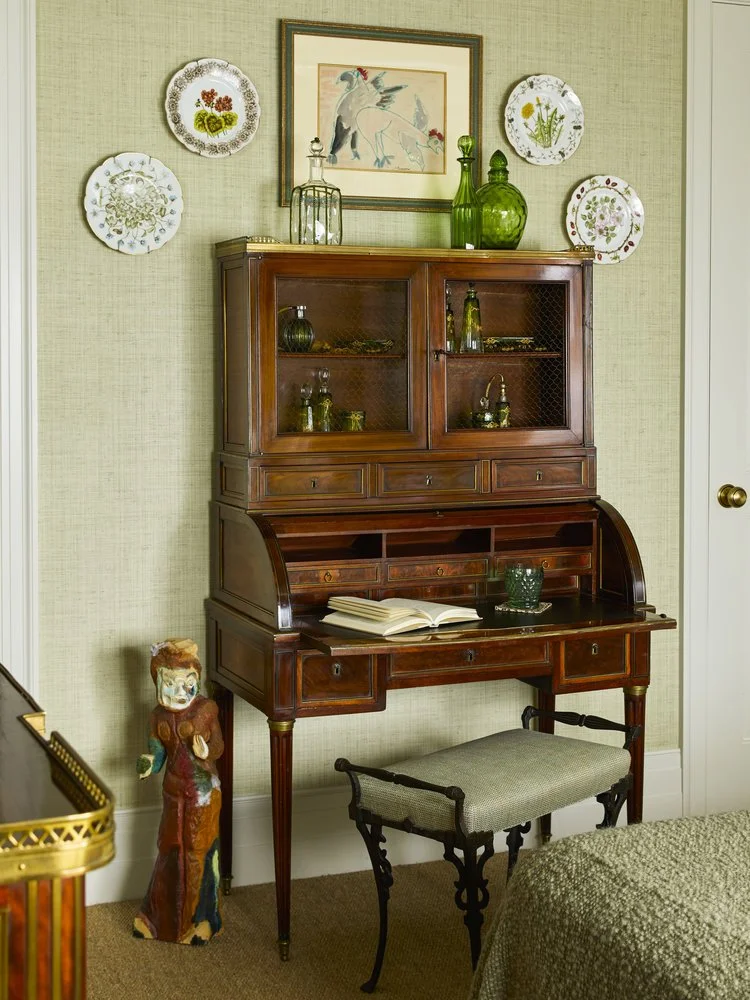 A vintage roll-top desk with an open book on it is positioned against a wall. Above it is a framed illustration of two birds, surrounded by decorative plates. Green glass bottles are displayed on top of the desk. A small upholstered stool is in front