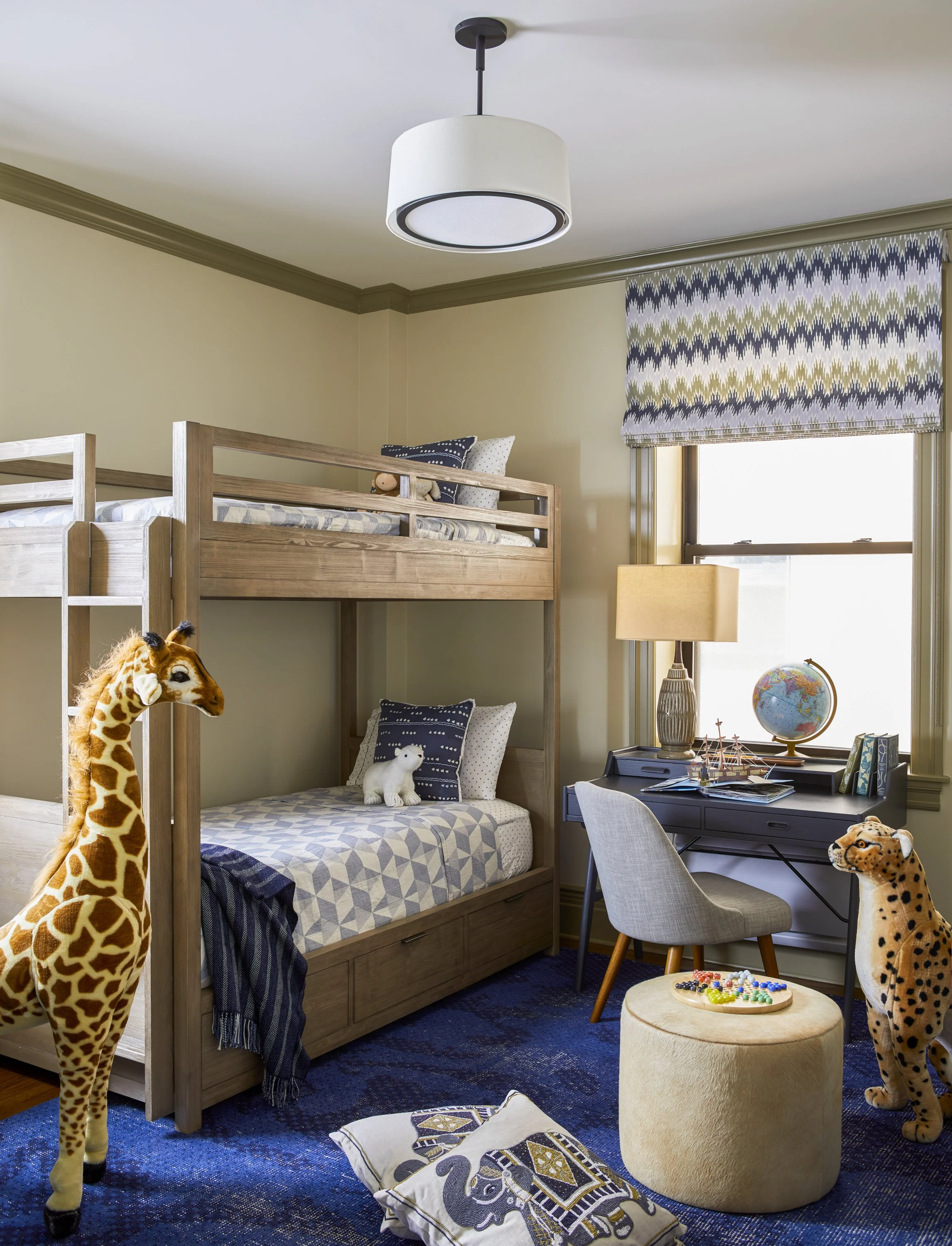 A cozy children's bedroom with a wooden bunk bed and colorful bedding, featuring plush giraffe and leopard toys. A study area with a blue desk, chair, globe, and books is near the window. The room has a blue textured rug, a ceiling light, and decorat