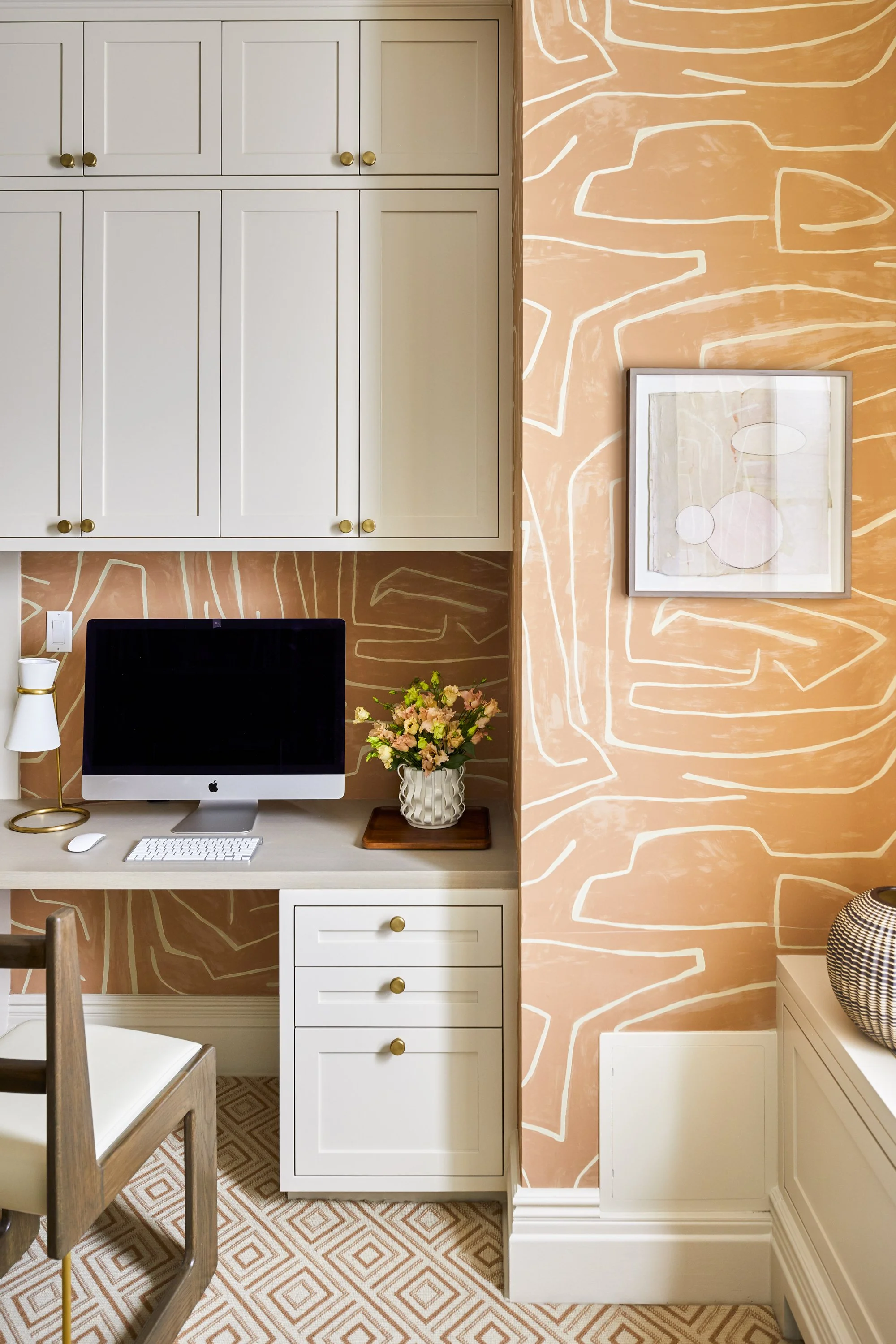 Home office space with white cabinets, a computer on a desk, a decorative lamp, flowers in a vase, geometric wallpaper, and patterned carpeting.