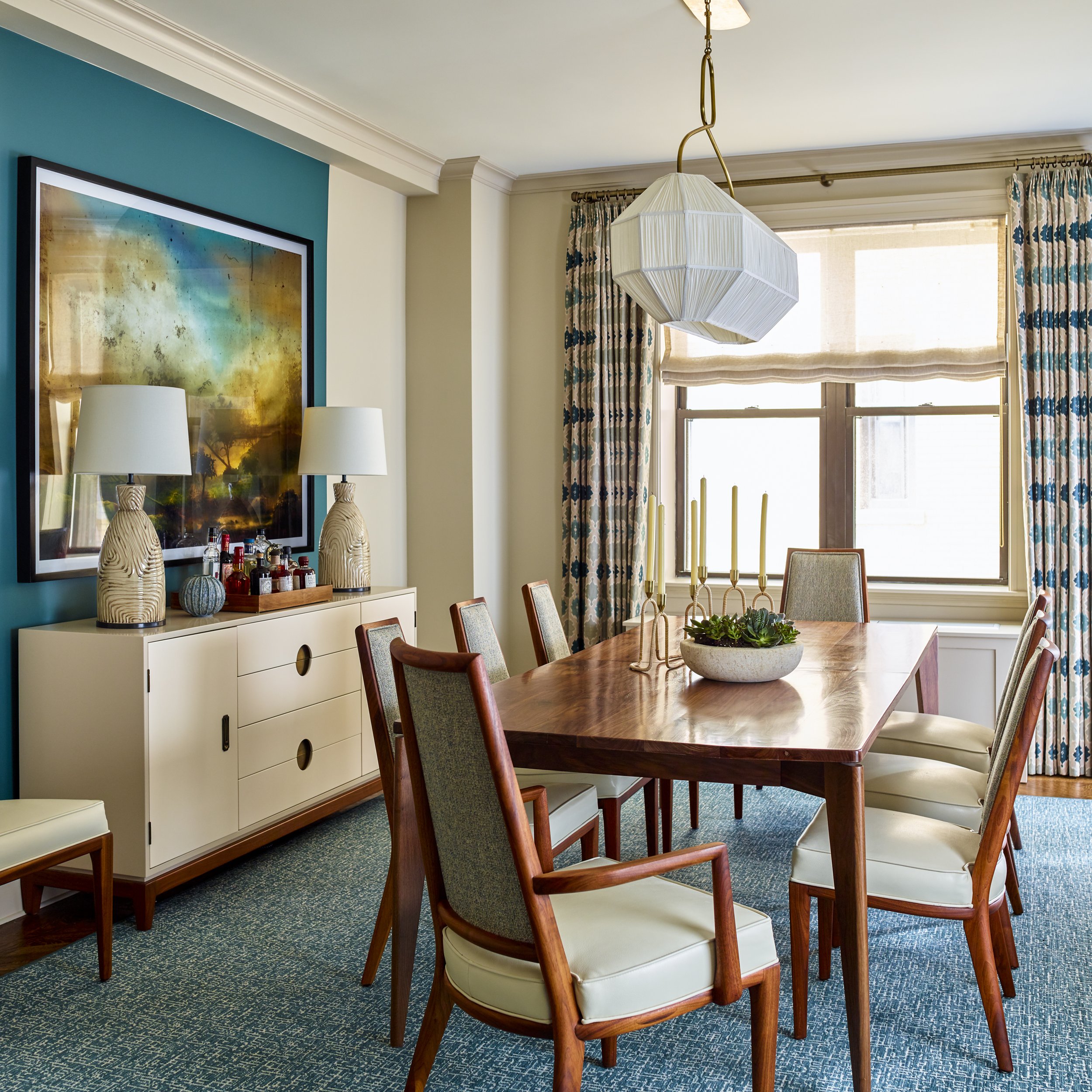 A modern dining room with a wooden table surrounded by six chairs. The room features a sideboard with two lamps and decorative items. A large framed artwork hangs on a teal accent wall. A geometric light fixture is centered above the table, which is 