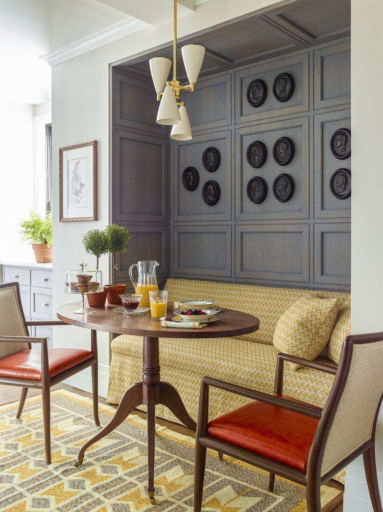 Cozy dining nook with round wooden table and two chairs, featuring a built-in cushioned bench with geometric pattern. Wall panels are adorned with black medallion decorations. A modern light fixture hangs above. Table set with a pitcher, juice, and b