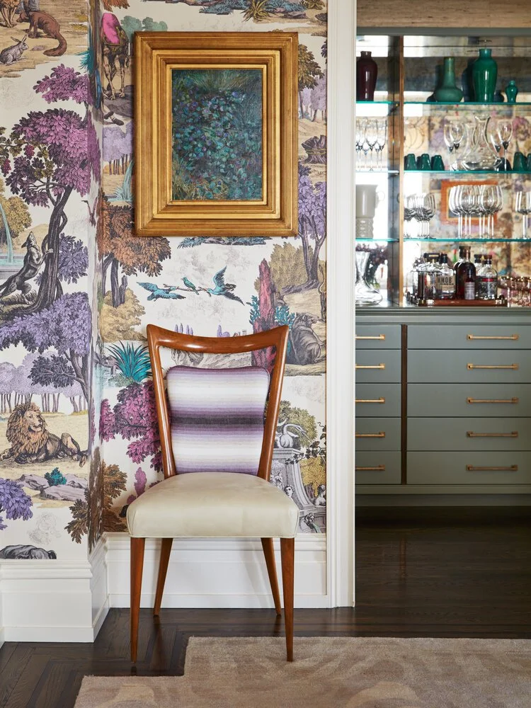 Elegant interior with a chair and colorful wallpaper, featuring nature motifs. A framed painting hangs above the chair, adjacent to a mirrored cabinet with glassware and bottles.