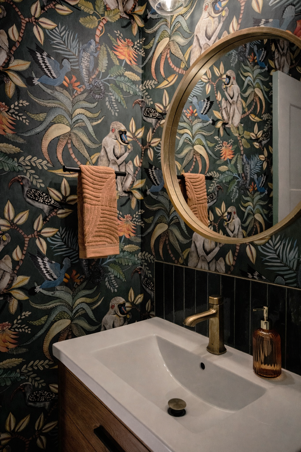 Bathroom with tropical animal-themed wallpaper featuring birds, snakes, and monkeys, round mirror, beige towel, sink with gold faucet, brown soap dispenser, and black tiled wall.