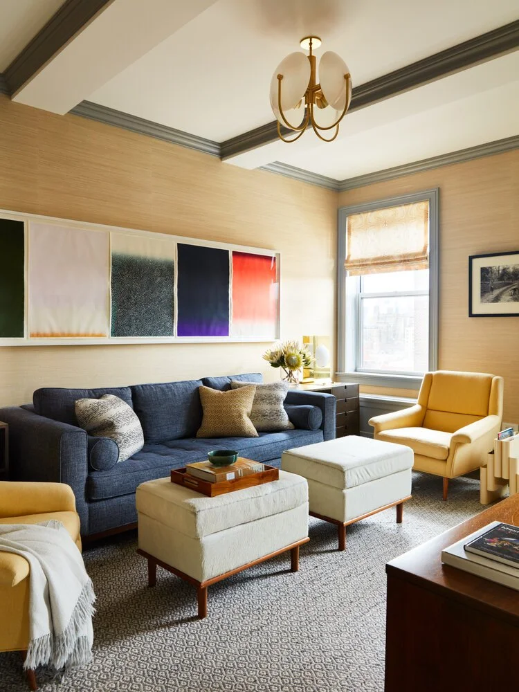 Cozy living room with a dark blue sofa, yellow armchairs, white ottomans, modern chandelier, abstract wall art, and a window with a beige shade.