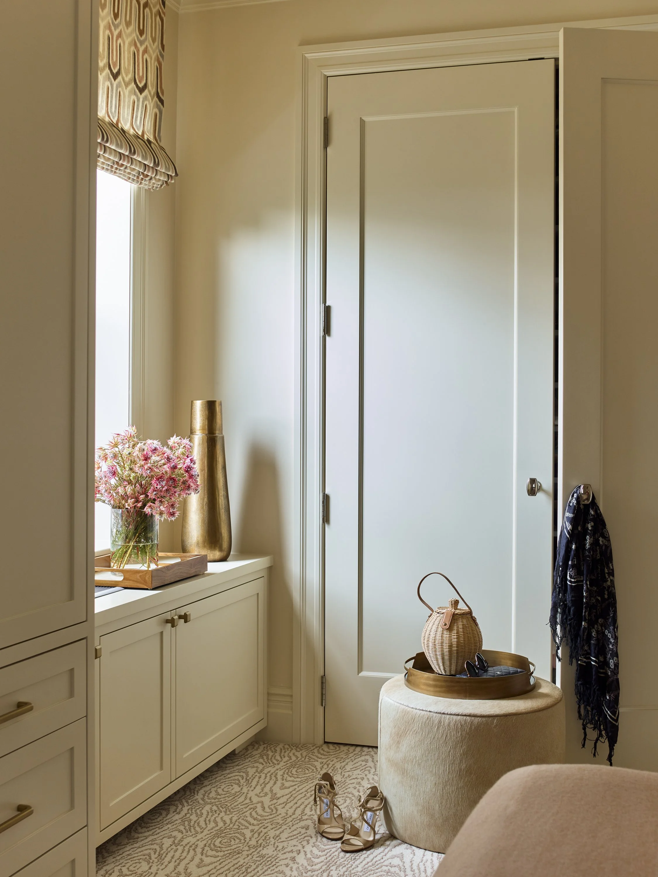 Interior room with beige decor, featuring a closed door, a cabinet with flowers in a vase, a tall brass vase, an ottoman with a circular tray, a wicker bag, sandals on the floor, and a patterned window shade.